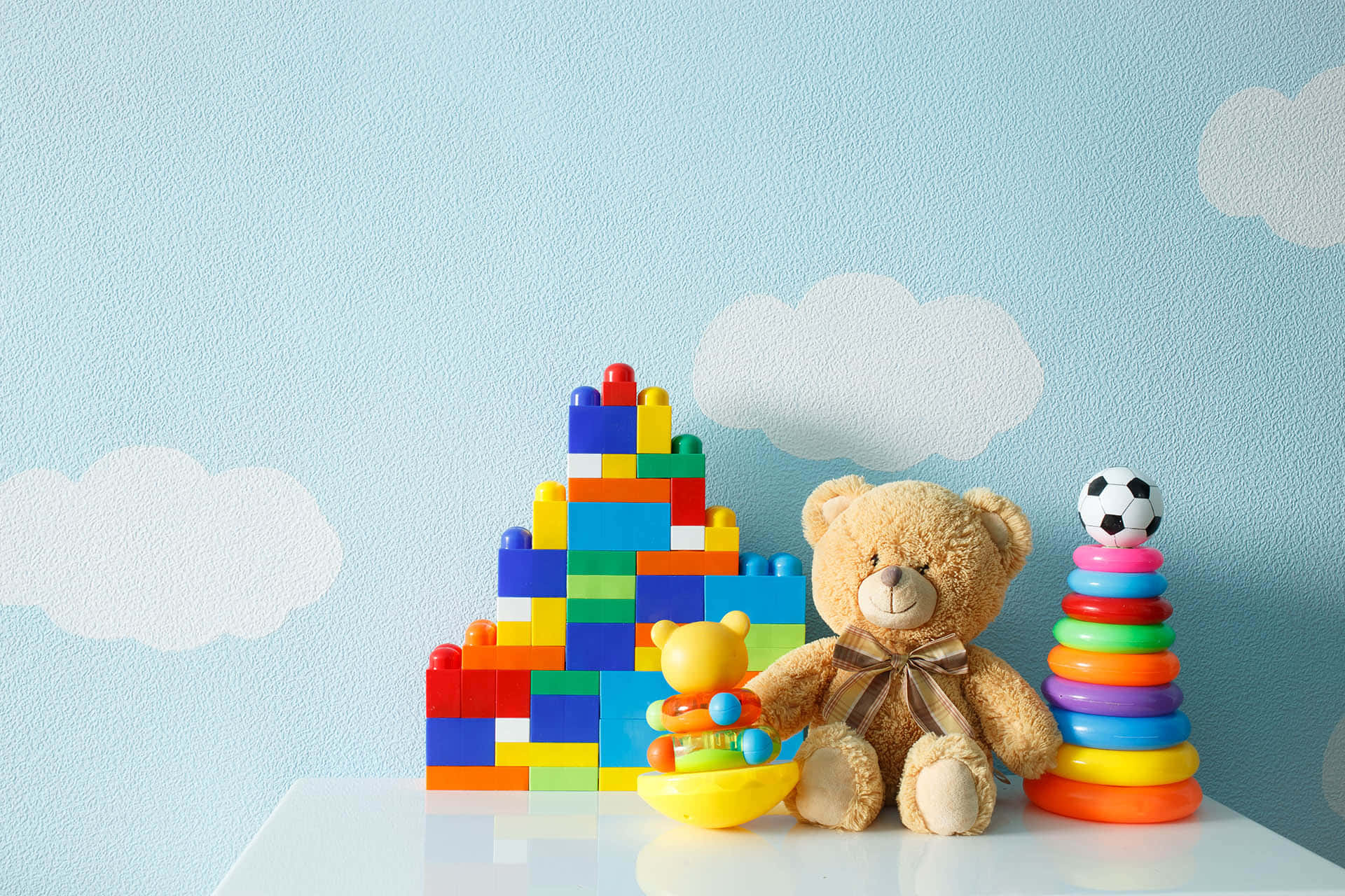 toys 桌布 壁紙 — 一隻泰迪熊坐在白色表面上，旁邊有色彩繽紛的積木和彩虹疊疊圈玩具，背景是淺藍色牆壁，上面畫有雲朵。