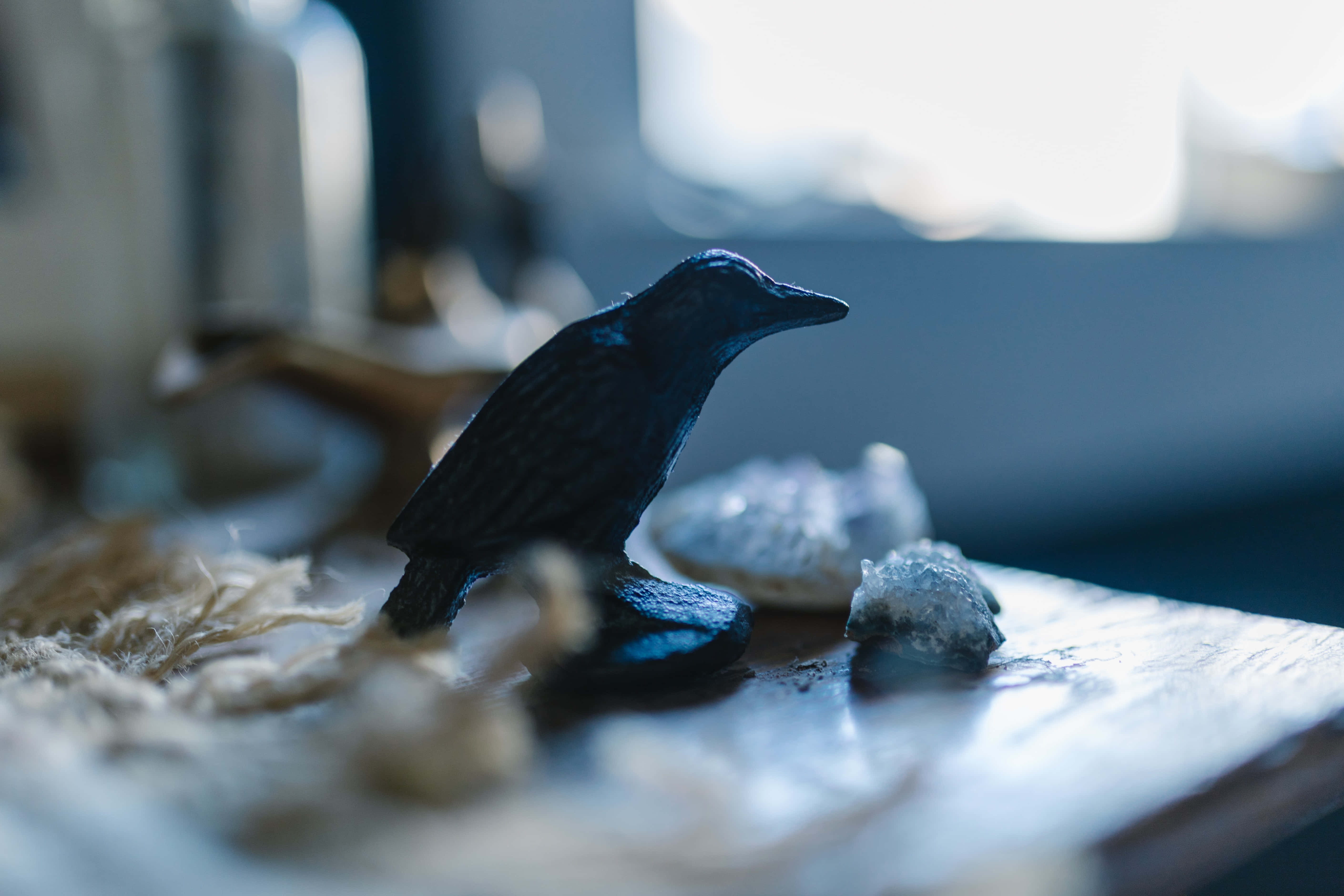 Raven 桌布 壁紙 — 一隻深藍色小鳥雕像棲息在木質表面上，旁邊有水晶狀物體和乾草，在柔和自然光下。
