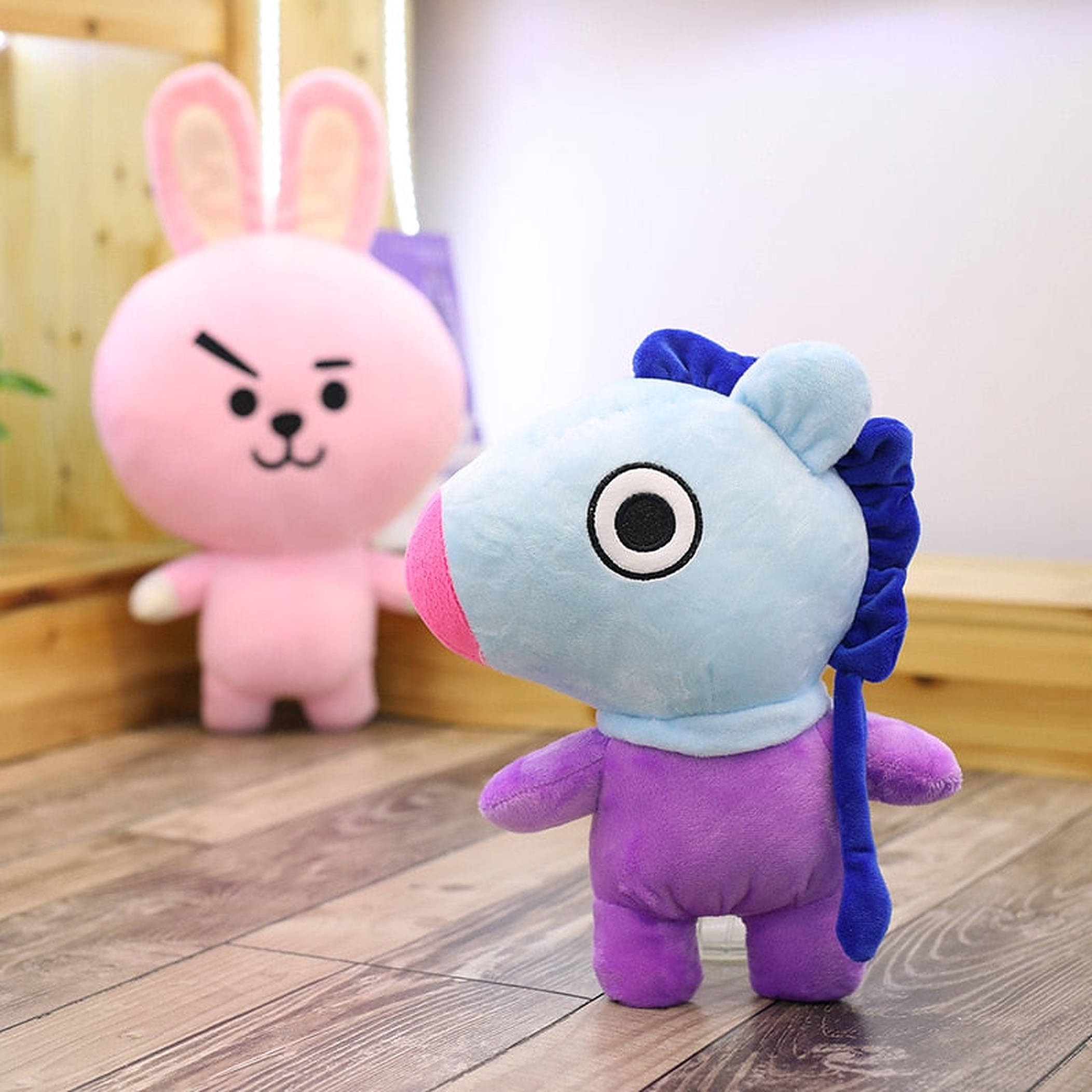 mang bt21 桌布 壁紙 — 一隻淺藍色毛絨獨角獸，有一隻大眼睛和紫色身體，站在木地板上，旁邊有一隻粉紅色毛絨兔玩具。