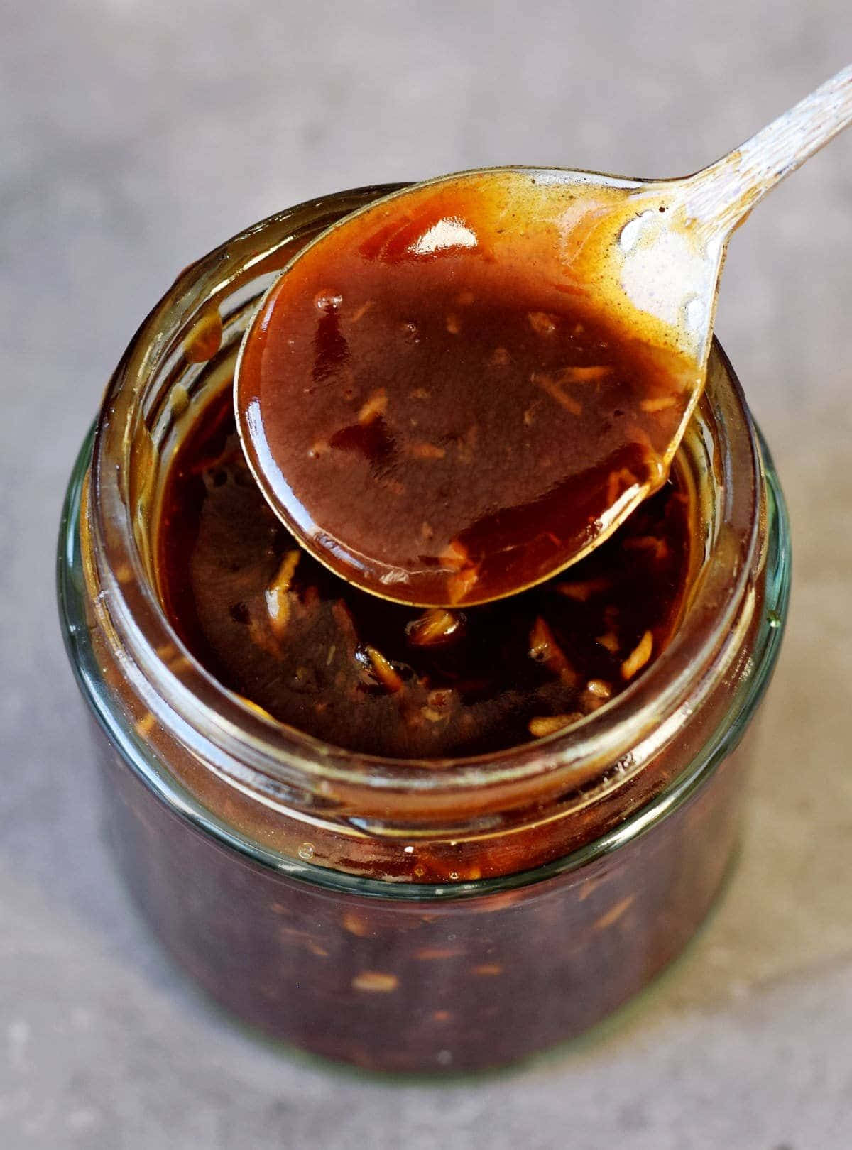 Szechuan Sauce 桌布 壁紙 — 一隻湯匙從玻璃罐中舀起帶有辣椒片的亮面深棕色醬汁，背景為中性灰色。