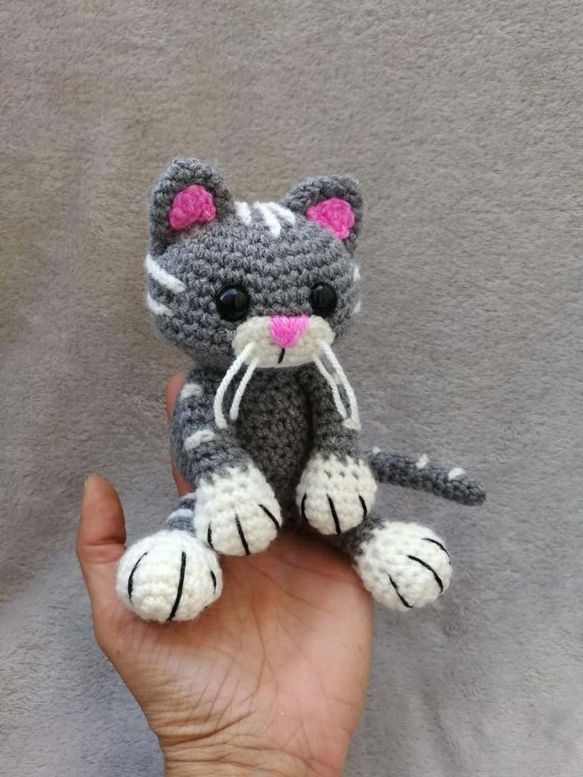 Crochet 桌布 壁紙 — 一隻灰白相間的小鉤針貓咪，有粉紅色的耳朵和鼻子，坐在一個人張開的手掌中，背景是柔和的灰色。