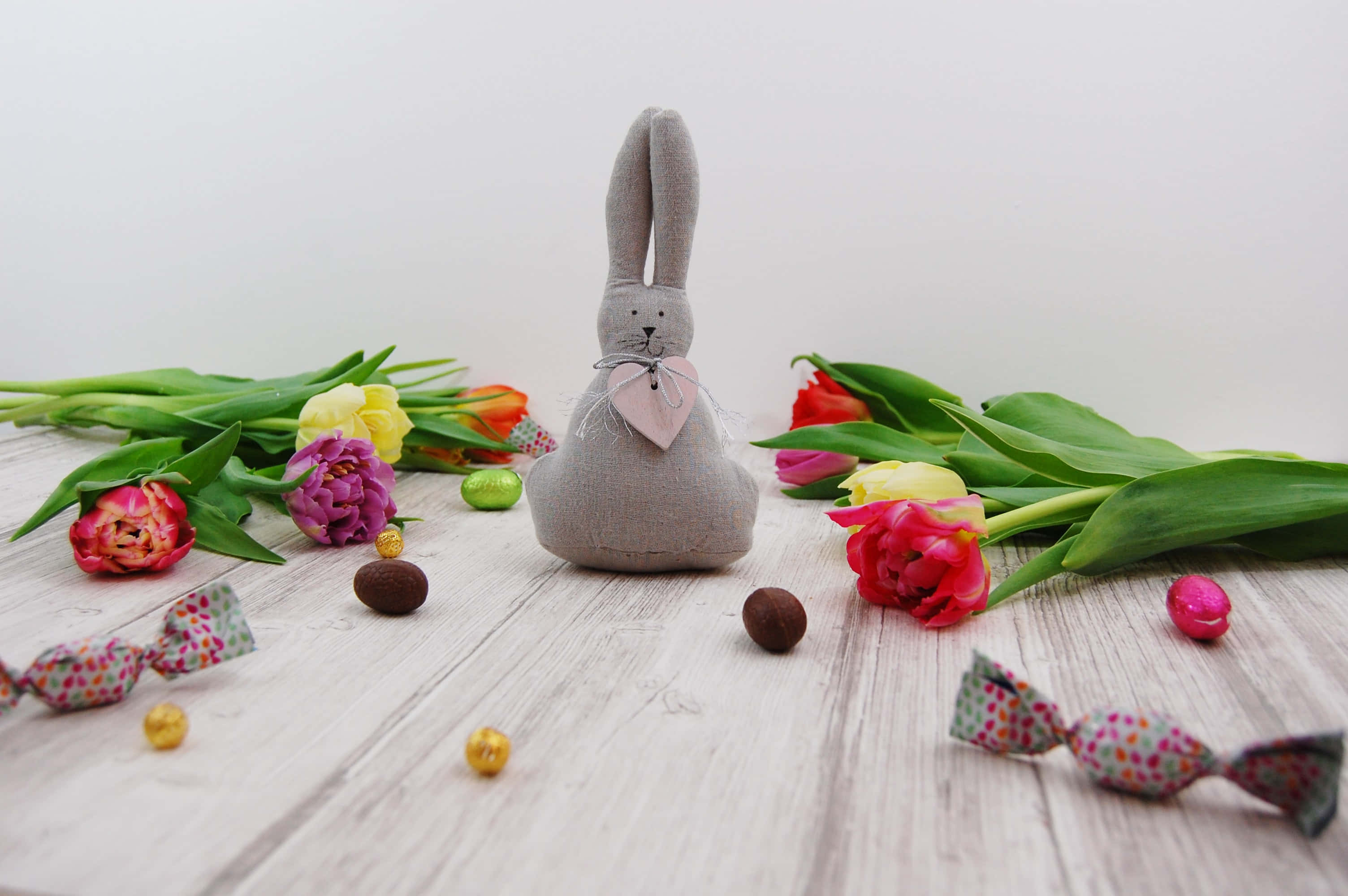 Easter Bunny 桌布 壁紙 — 一隻灰色布兔戴著粉紅色蝴蝶結，坐在白色粉刷的木質表面上，周圍環繞著色彩繽紛的鬱金香和包裝糖果。