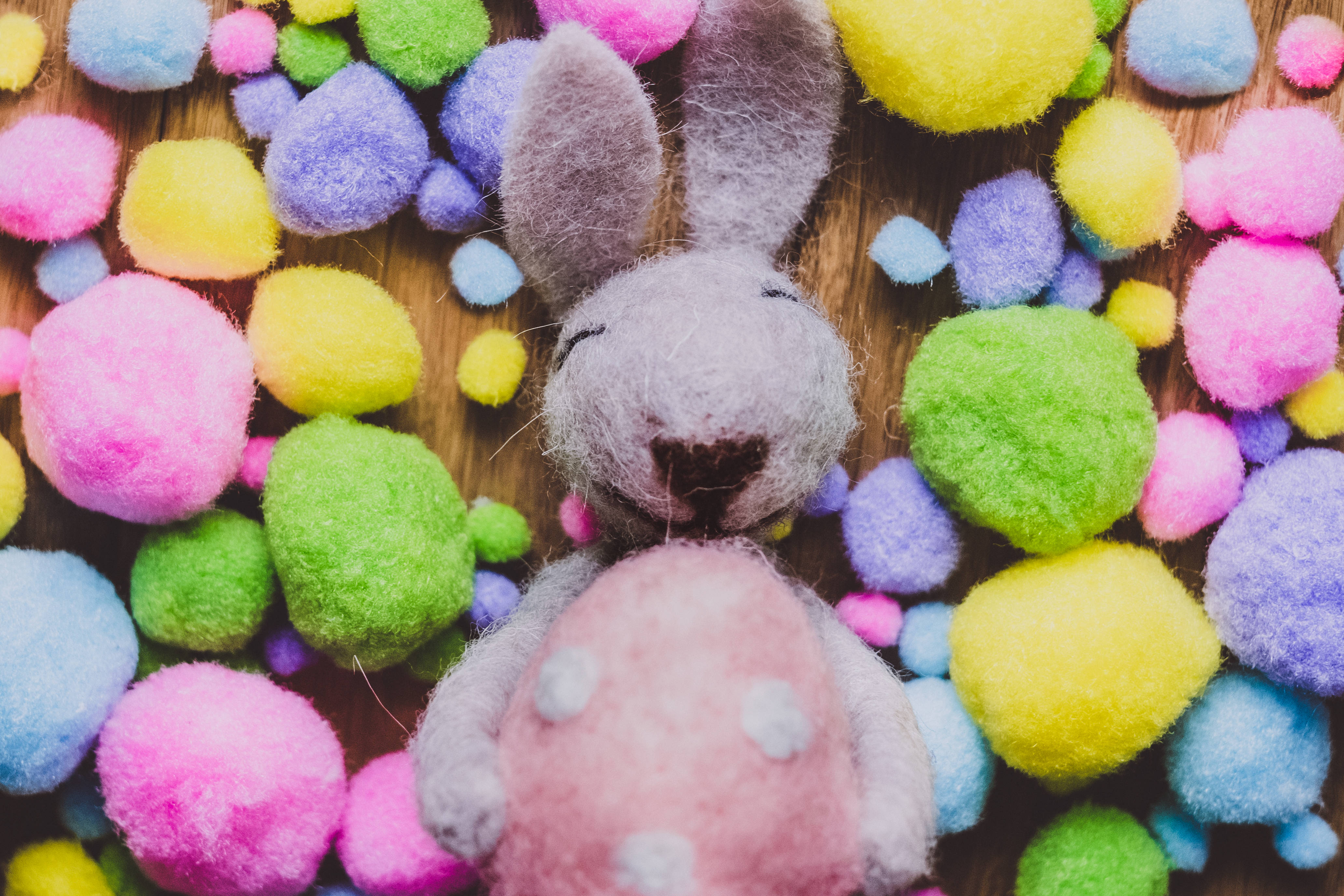 Happy Easter 桌布 壁紙 — 一隻灰色氈兔拿著一個粉紅色圓點蛋，周圍是色彩繽紛的毛毛球在木頭上。