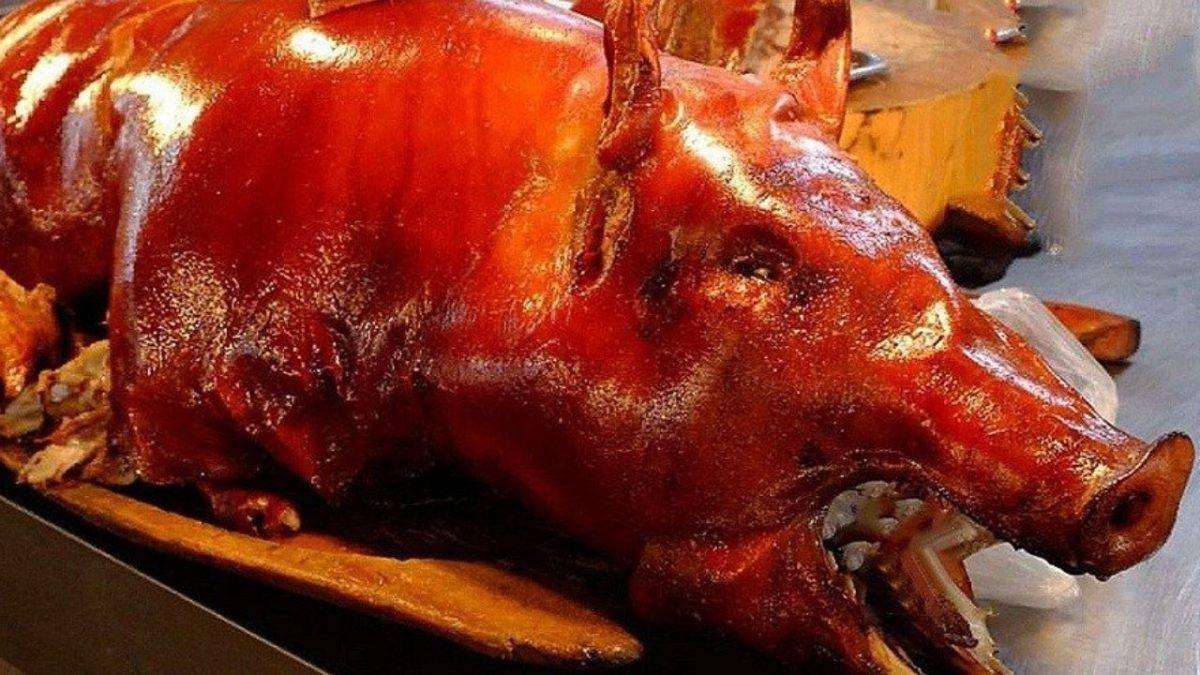 Pig 桌布 壁紙 — 一隻烤全豬，金黃酥脆的豬皮在明亮的燈光下閃閃發光，放在木製托盤上。