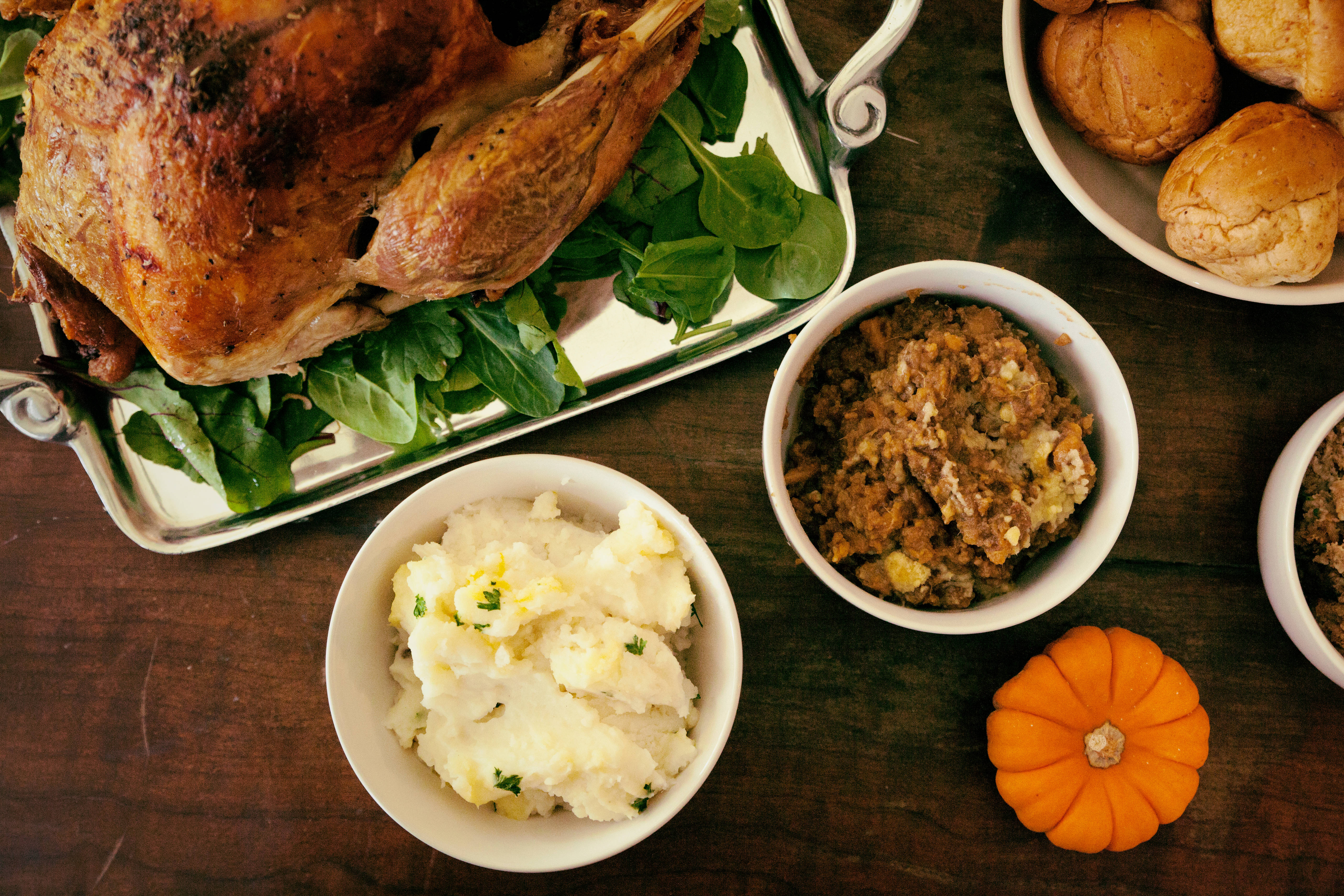 Happy Thanksgiving 桌布 壁紙 — 一隻烤火雞放在銀盤上的菠菜上，周圍是馬鈴薯泥、餡料、麵包捲和一個小橙色南瓜，放在深色木桌上。