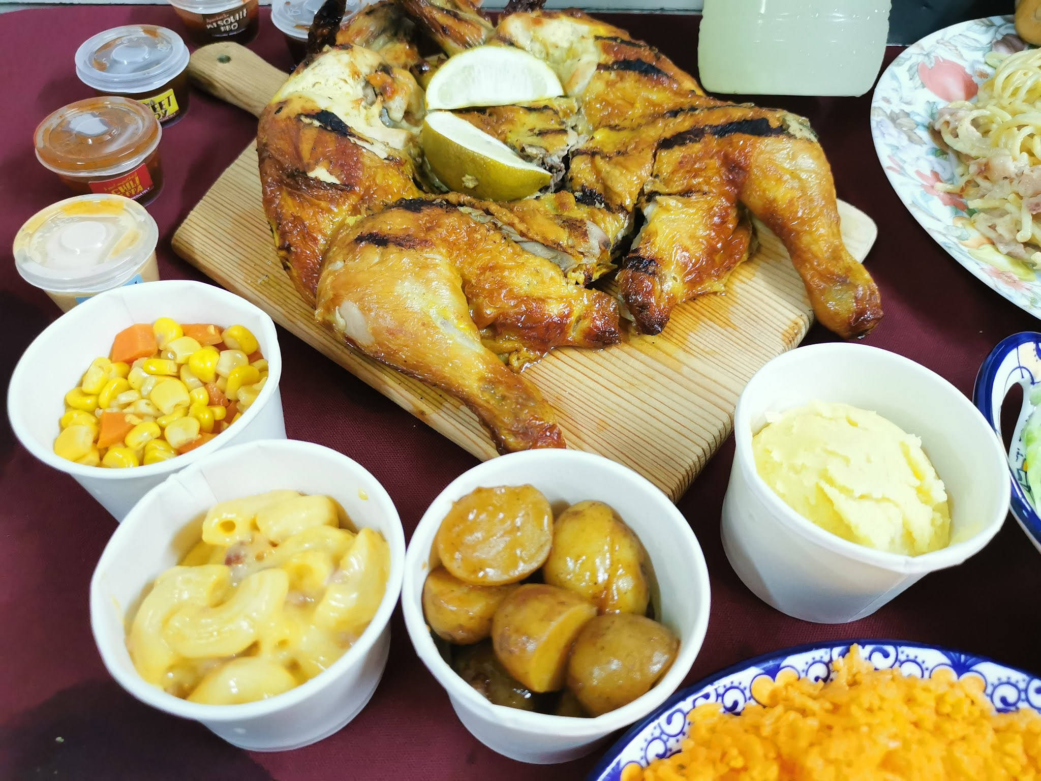 Delicious Food 桌布 壁紙 — 一隻烤雞搭配萊姆角放在木板上，周圍有通心粉起司、玉米和馬鈴薯等配菜，置於栗色桌布上。