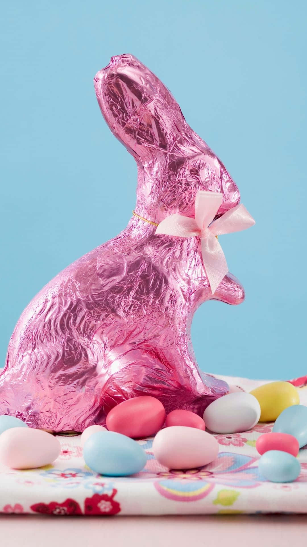 Easter Bunny 桌布 壁紙 — 一隻用粉紅色箔紙包裹、帶有蝴蝶結的大型復活節巧克力兔，坐在花卉布上，周圍是粉彩糖果蛋，背景是淺藍色。