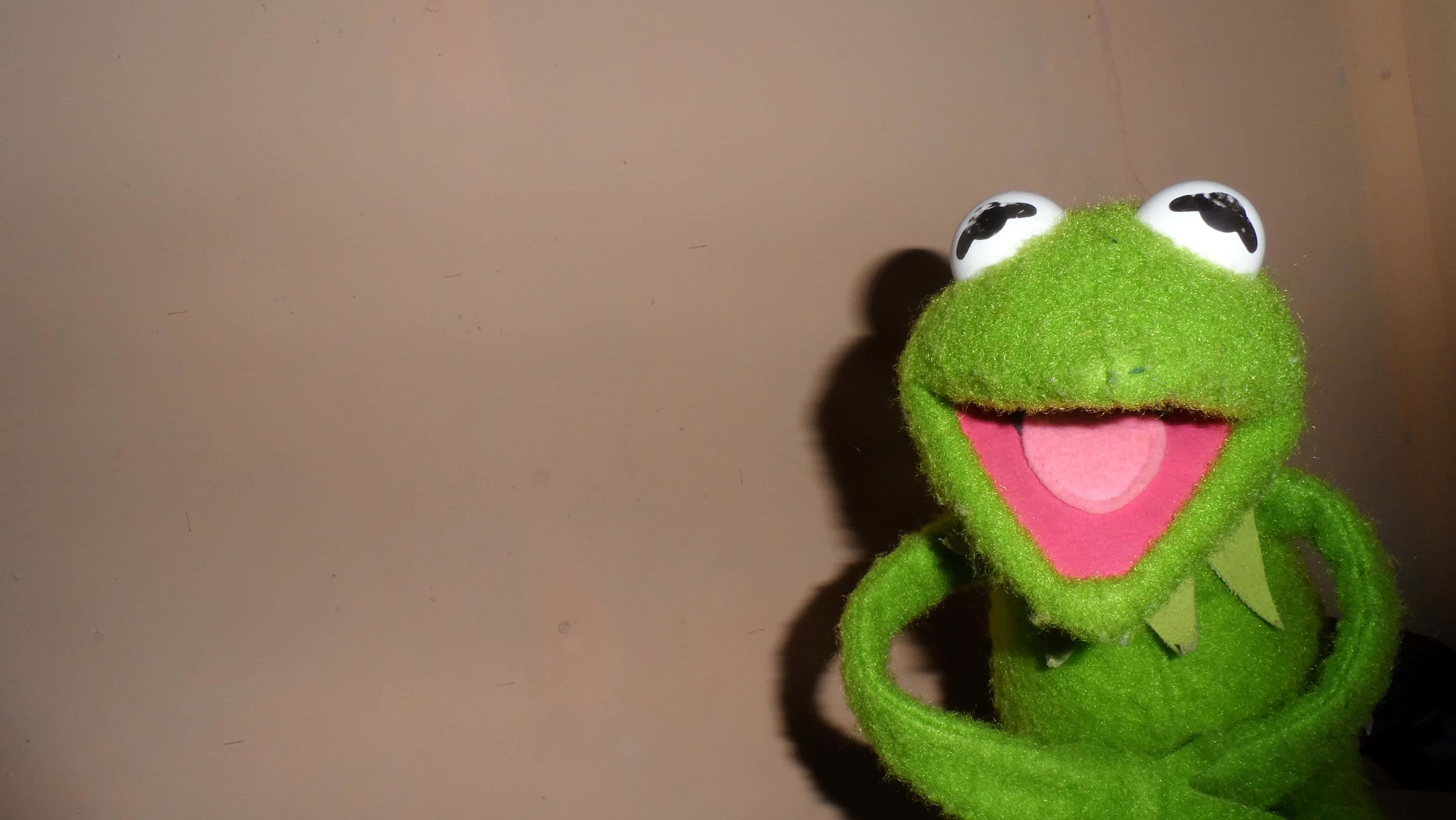 Aesthetic Frog 桌布 壁紙 — 一隻用綠色絨布製成、表情歡樂的青蛙手偶，有著白色大眼睛和寬闊的粉紅色嘴巴，對著純色米色牆壁微笑。