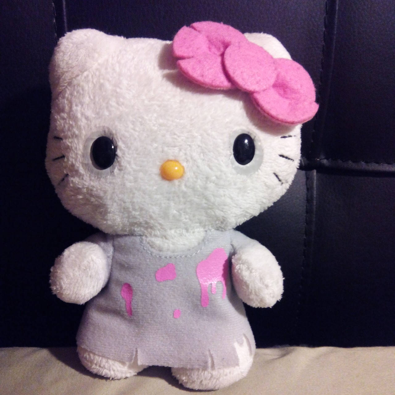 Hello Kitty Halloween 桌布 壁紙 — 一隻白色Hello Kitty絨毛玩偶，戴著粉紅色蝴蝶結，灰色裙子上有粉紅色油漆噴濺，坐在深色皮革背景前。
