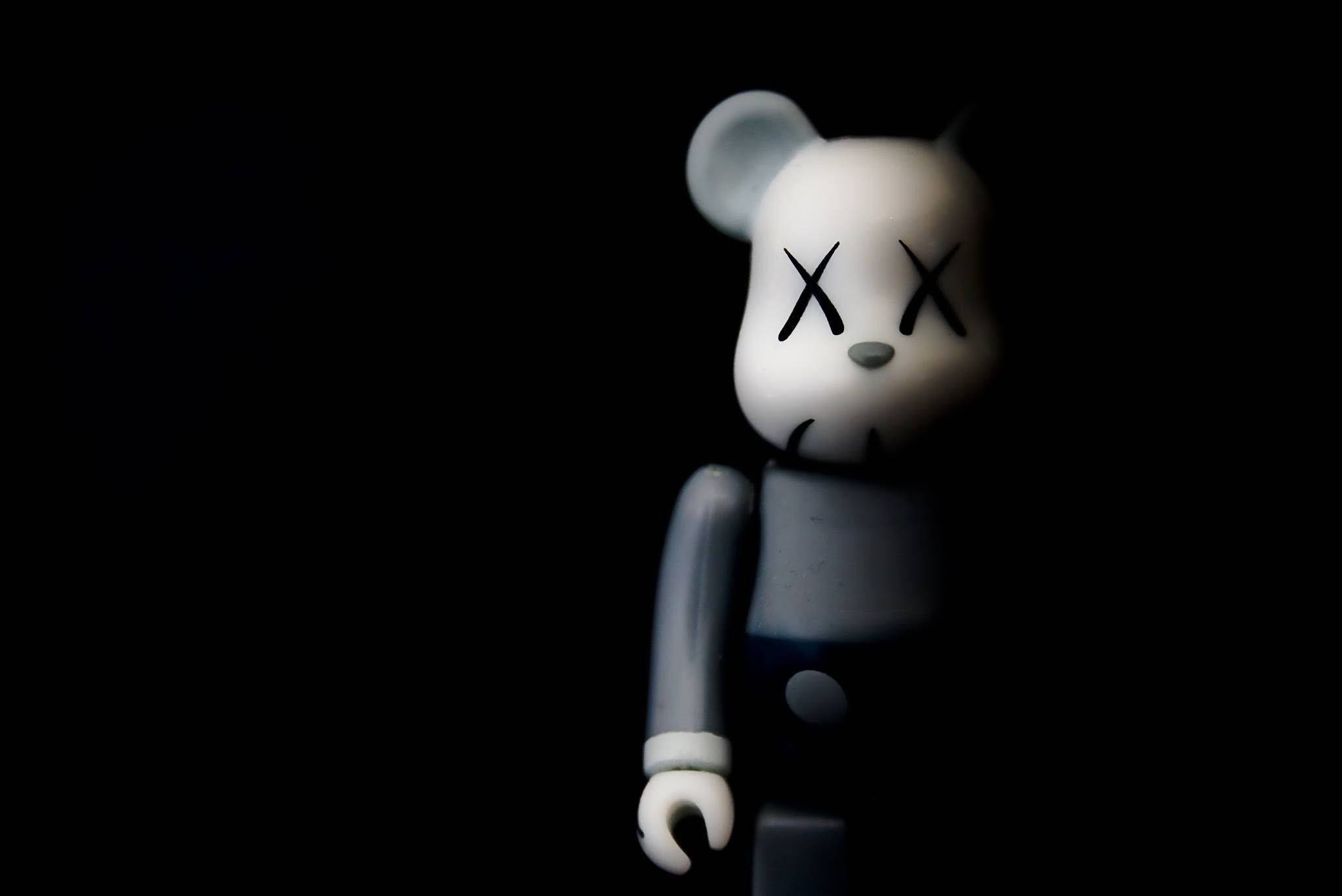 kaws 4K 桌布 壁紙 — 一隻白色KAWS熊公仔，帶有X形眼睛，在昏暗光線下站在黑色背景前。