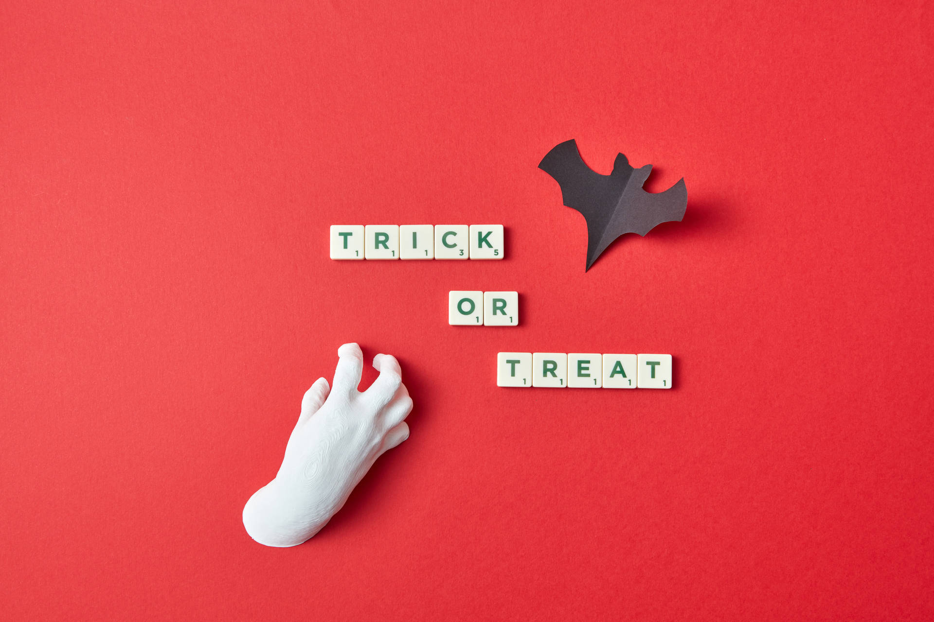 萬聖節動圖 桌布 壁紙 — 一隻白色塑膠手伸向拼出 trick or treat 的 Scrabble 字母磚，背景為大膽的紅色，並有一隻黑色紙蝙蝠。