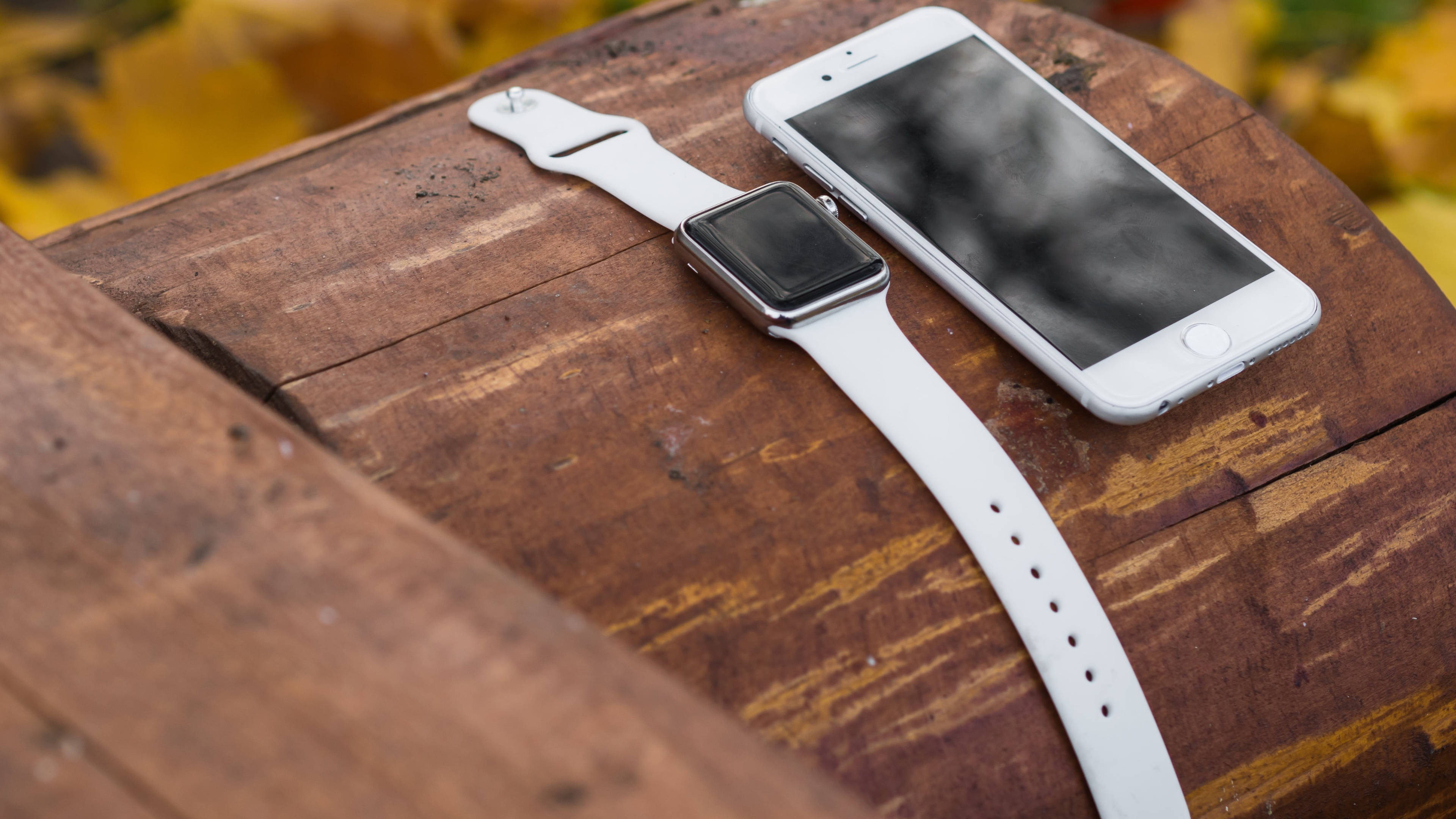Apple Watch 動態 桌布 壁紙 — 一隻白色智慧手錶和智慧型手機放在經風化的木質表面上，背景有模糊的秋葉。