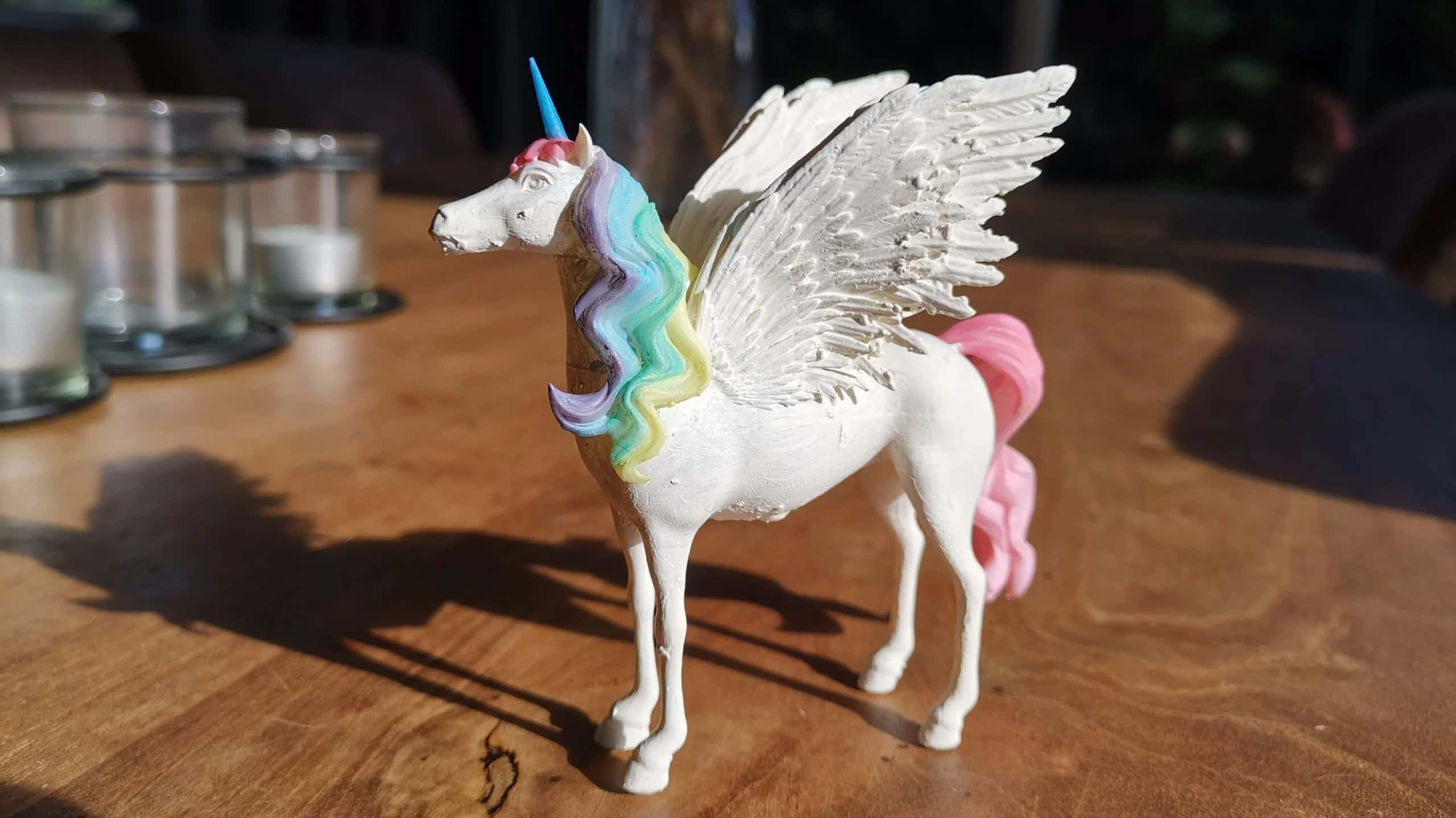 3d Unicorn 桌布 壁紙 — 一隻白色有翼獨角獸公仔，擁有彩虹鬃毛和粉紅色尾巴，站在陽光照射的木桌上。