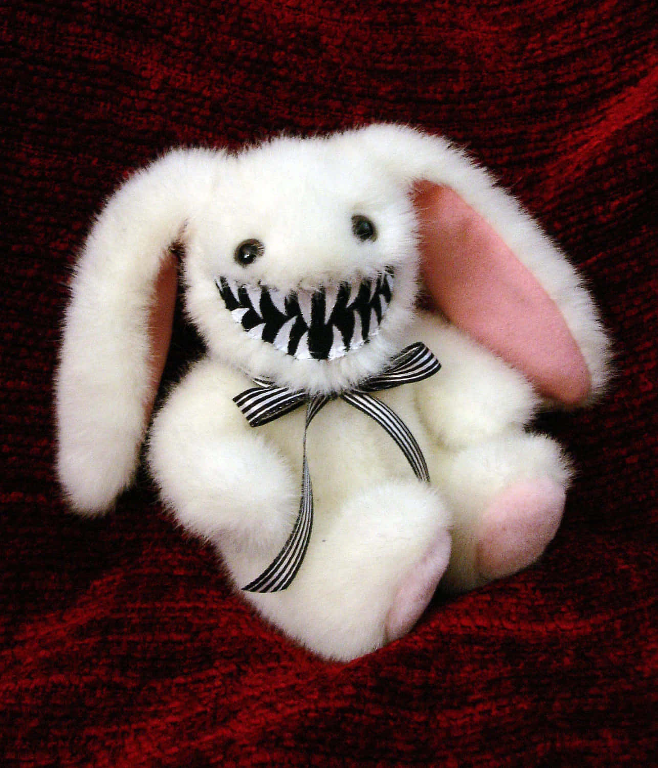 Creepy Easter Bunny 桌布 壁紙 — 一隻白色毛絨兔子，帶著寬闊的黑白條紋笑容，坐在深紅色紋理布料上。