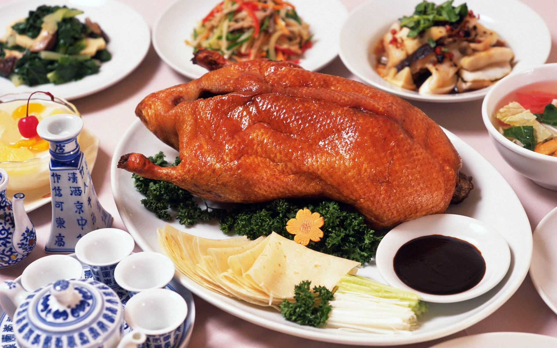 4K Food 桌布 壁紙 — 一隻皮光肉滑的全隻烤鴨坐在中央，周圍是色彩繽紛的配菜和傳統藍白瓷器茶具。