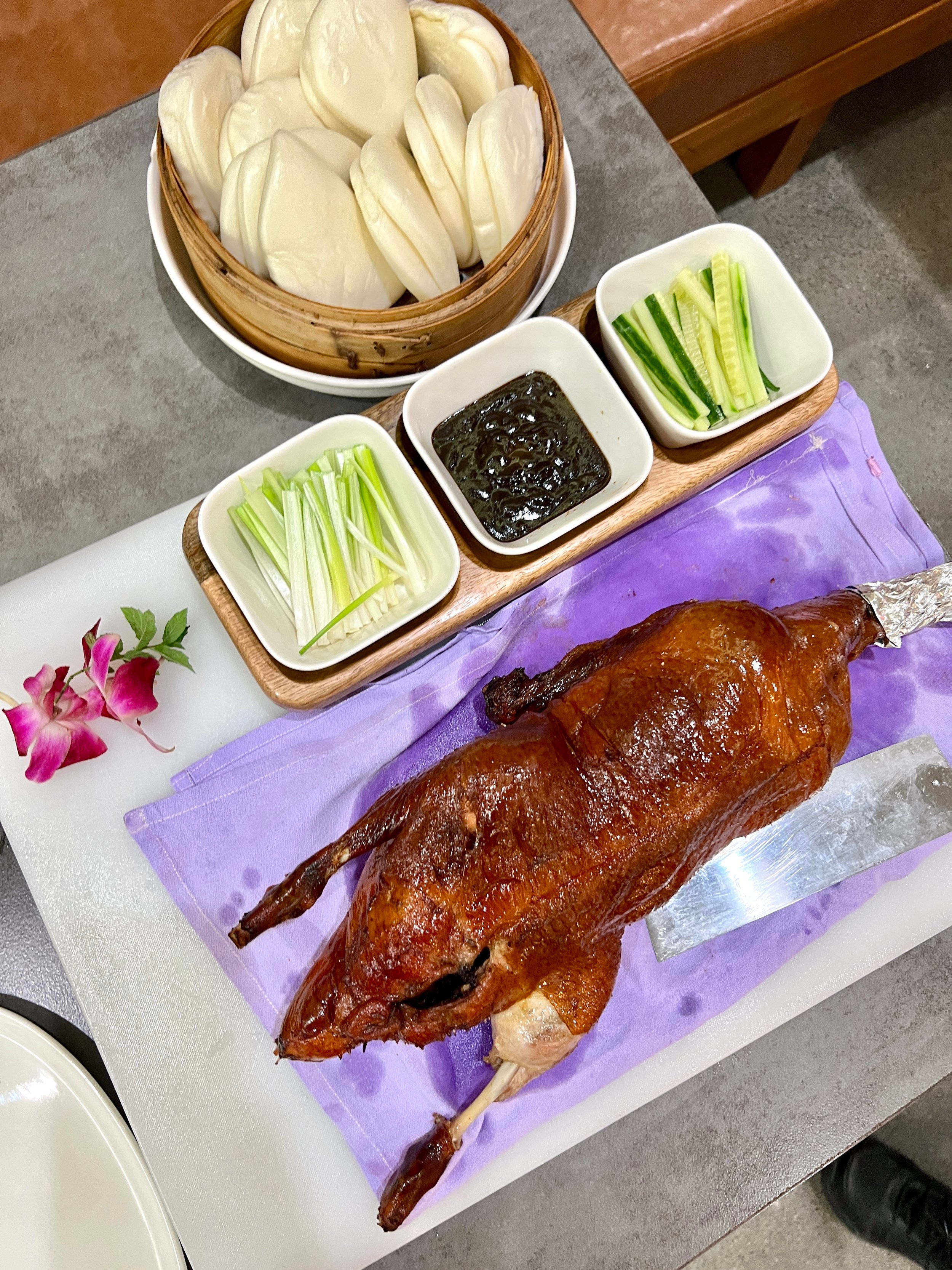 鴨子 桌布 壁紙 — 一隻皮光肉滑的整隻烤鴨放在紫色布上，旁邊有蒸包和黃瓜蔥花配菜。