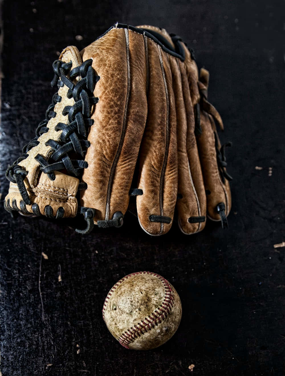 Baseball Gloves 桌布 壁紙 — 一隻磨損的棕色皮革棒球手套和一個有刮痕的棒球放在深色紋理表面上。