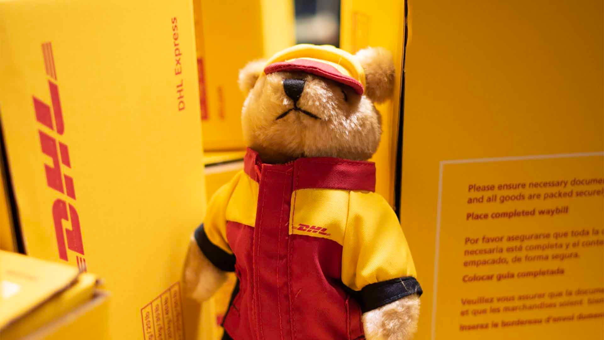Dhl 桌布 壁紙 — 一隻穿著DHL送貨制服的毛絨玩具熊站在黃色的送貨箱之間。