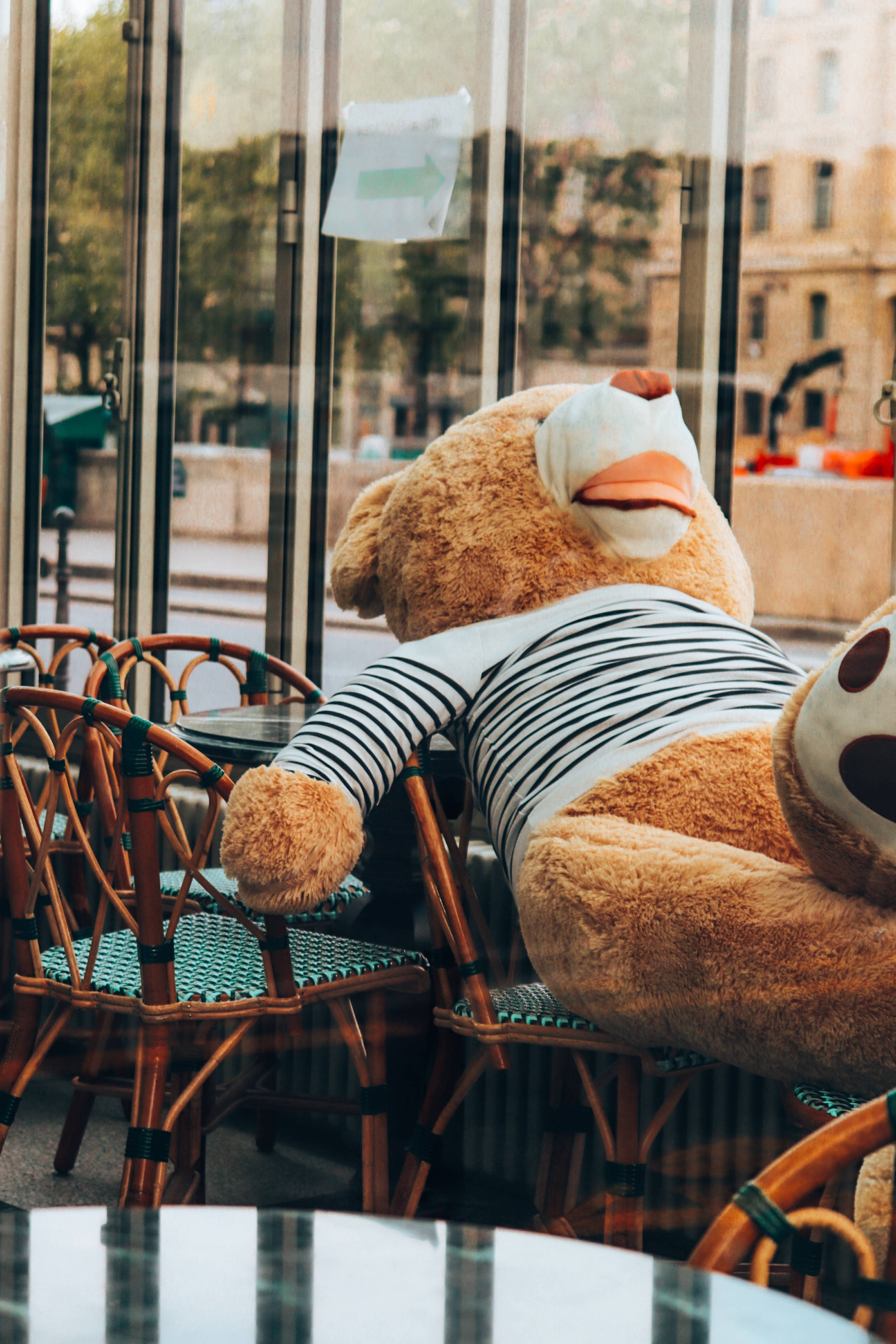Cute Bear 桌布 壁紙 — 一隻穿著條紋襯衫的巨大毛絨泰迪熊，懶洋洋地躺在咖啡館內的藤椅上，咖啡館的玻璃窗反射出城市街道的景象。