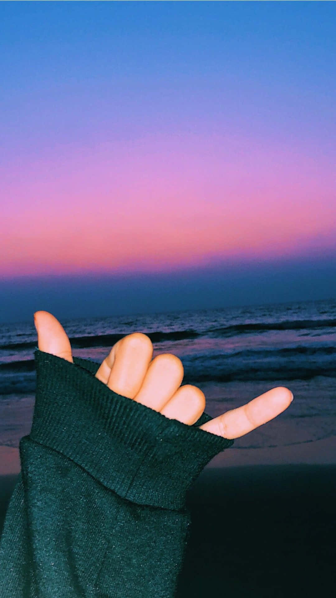 Aesthetic Summer 桌布 壁紙 — 一隻穿著深綠色毛衣的手，在海洋上充滿活力的紫色和藍色日落背景下做出沙卡手勢。