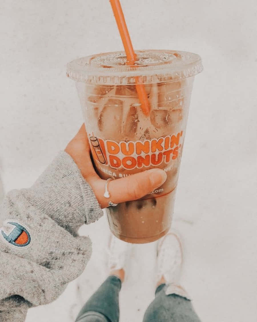 Dunkin Donuts 桌布 壁紙 — 一隻穿著灰色連帽衫的手，在雪景背景下拿著一杯帶有橙色吸管的Dunkin' Donuts冰咖啡。