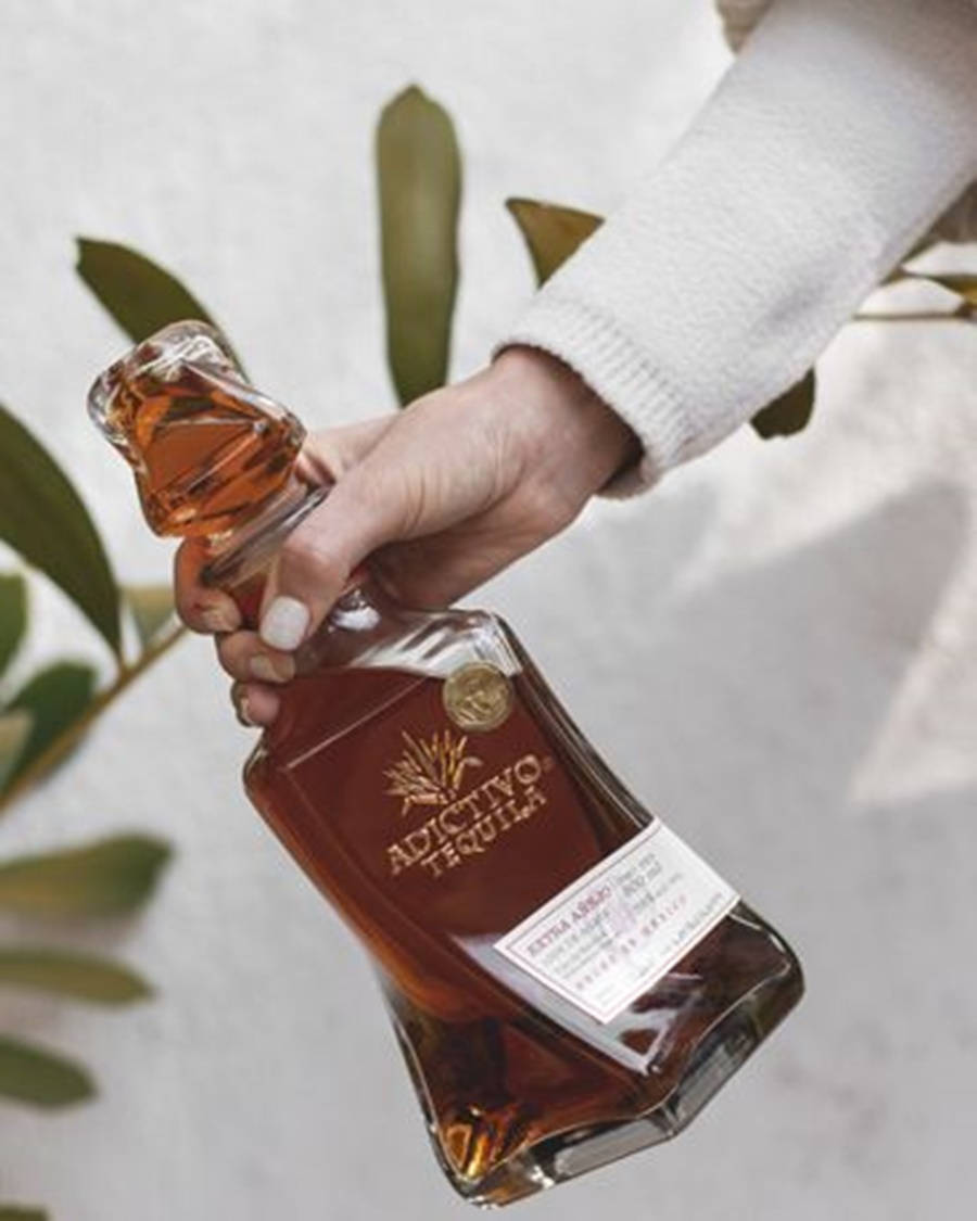 21:9 桌布 壁紙 — 一隻穿著白色毛衣的手，拿著一瓶Adictivo Tequila，背景是柔和的白色牆壁和綠葉。