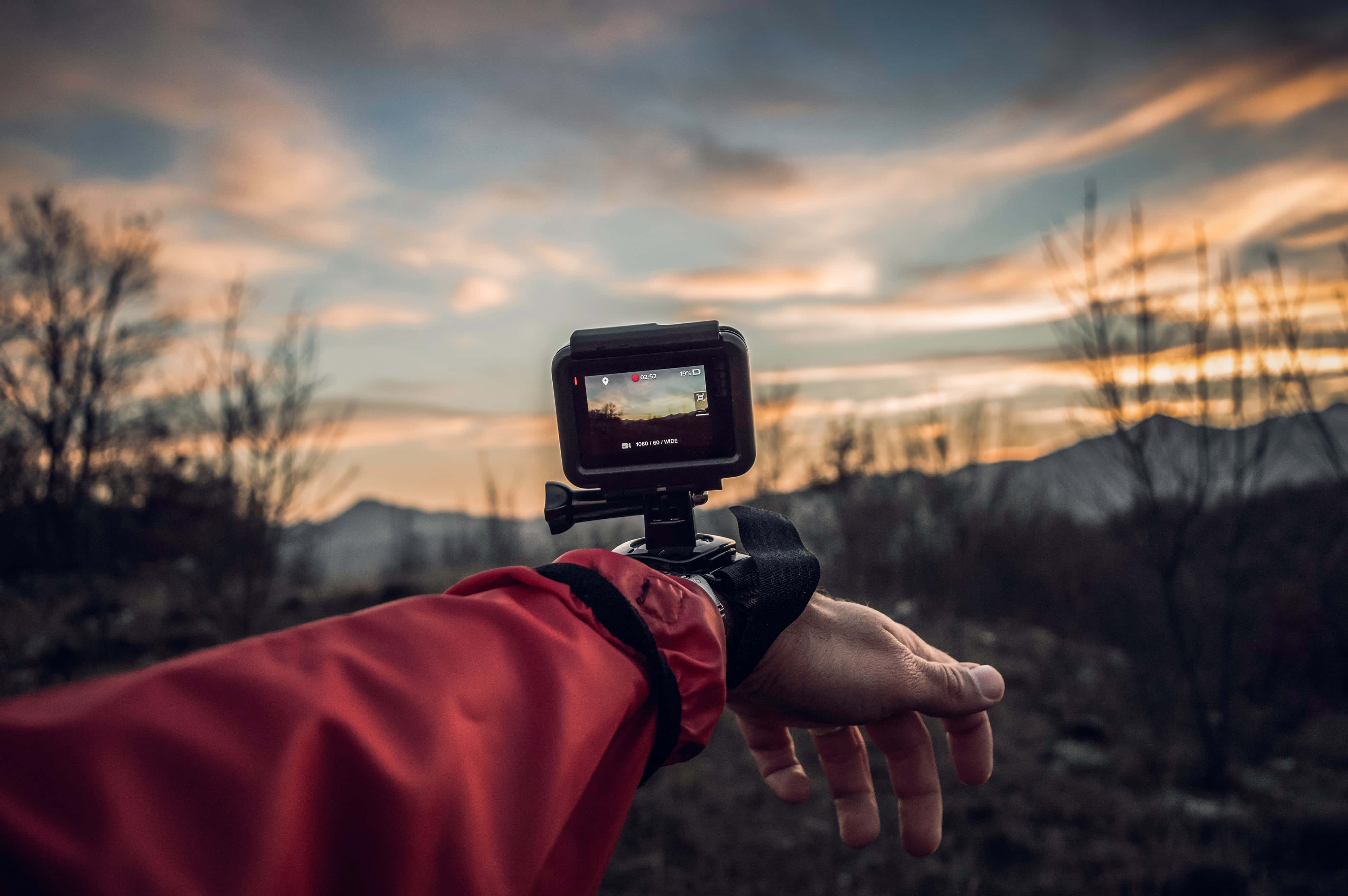gopro 桌布 壁紙 — 一隻穿著紅色外套的手握著手腕式運動相機，捕捉遠山和光禿樹木上戲劇性的日落。