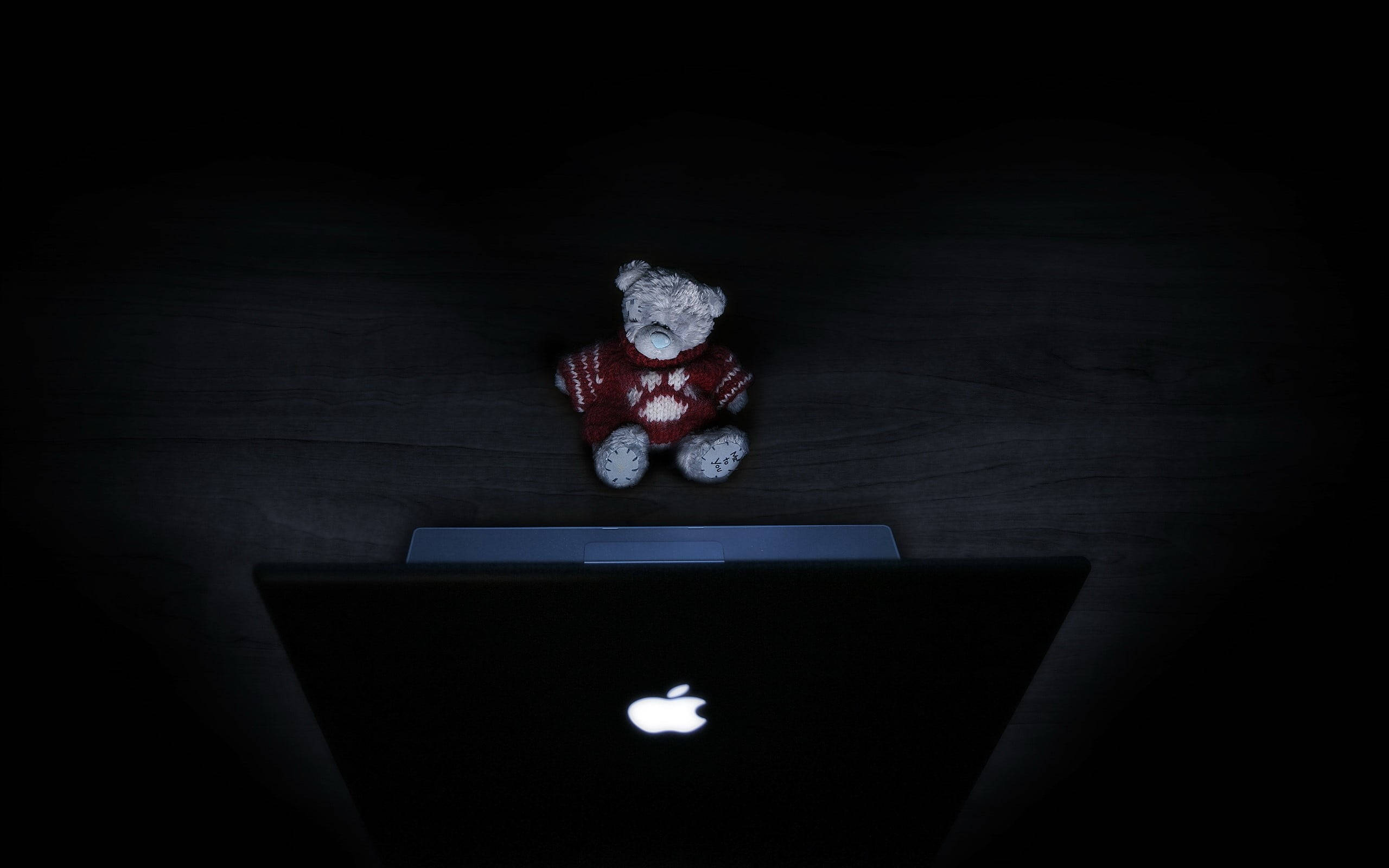 MacBook 動態圖片 桌布 壁紙 — 一隻穿著紅色毛衣的小熊坐在黑暗房間中關閉的筆記型電腦上，被發光的Apple標誌照亮。