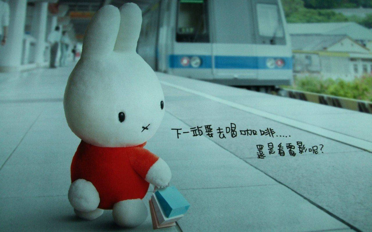 米飛兔 桌布 壁紙 — 一隻穿著紅色襯衫的白色兔毛公仔坐在火車月台上，拿著一本書，背景中有一列火車正在駛近。