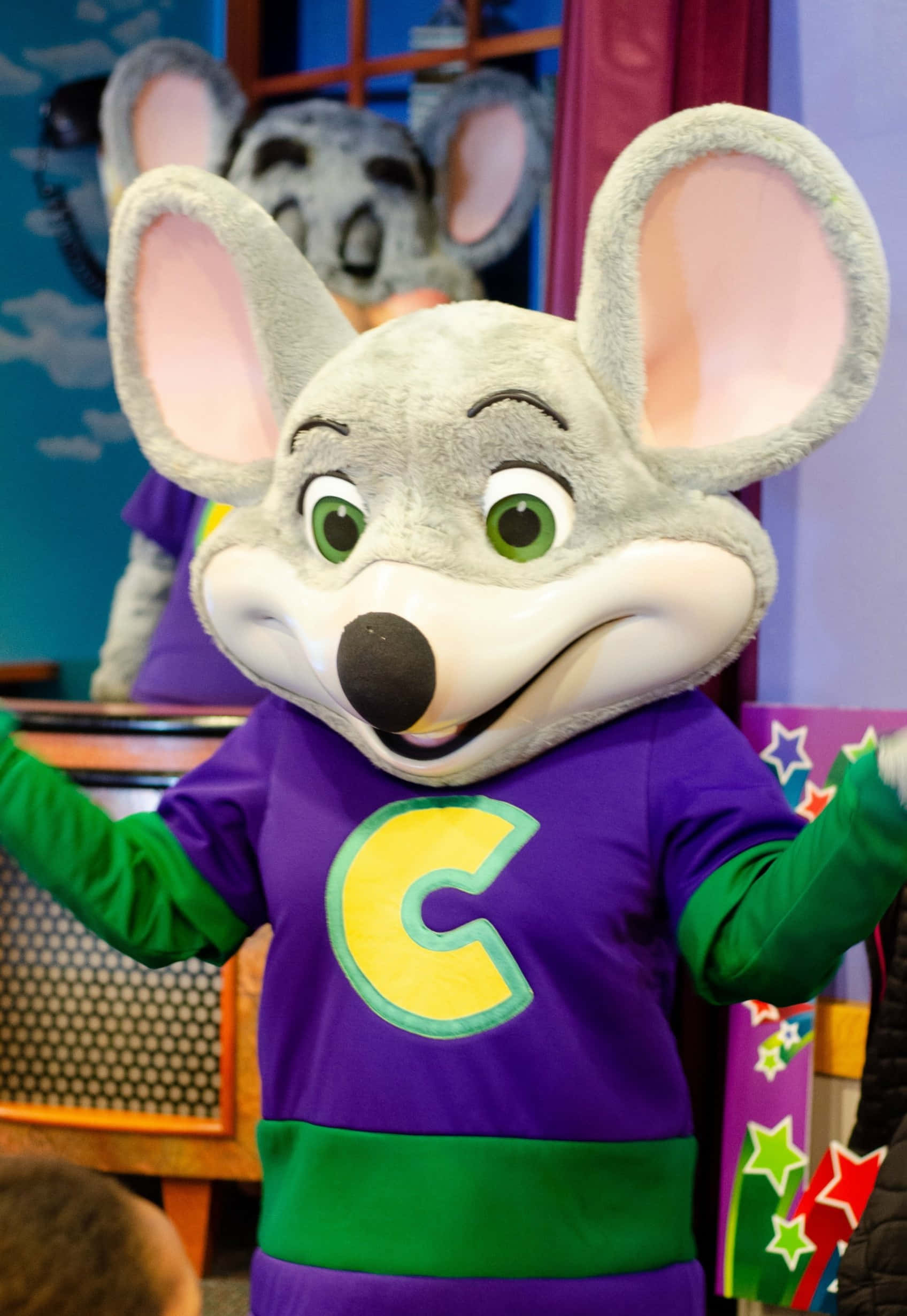 Chuck E Cheese 桌布 壁紙 — 一隻穿著紫綠色襯衫、帶有黃色C字的開心灰色老鼠吉祥物，在色彩繽紛的室內場景中燦爛微笑。