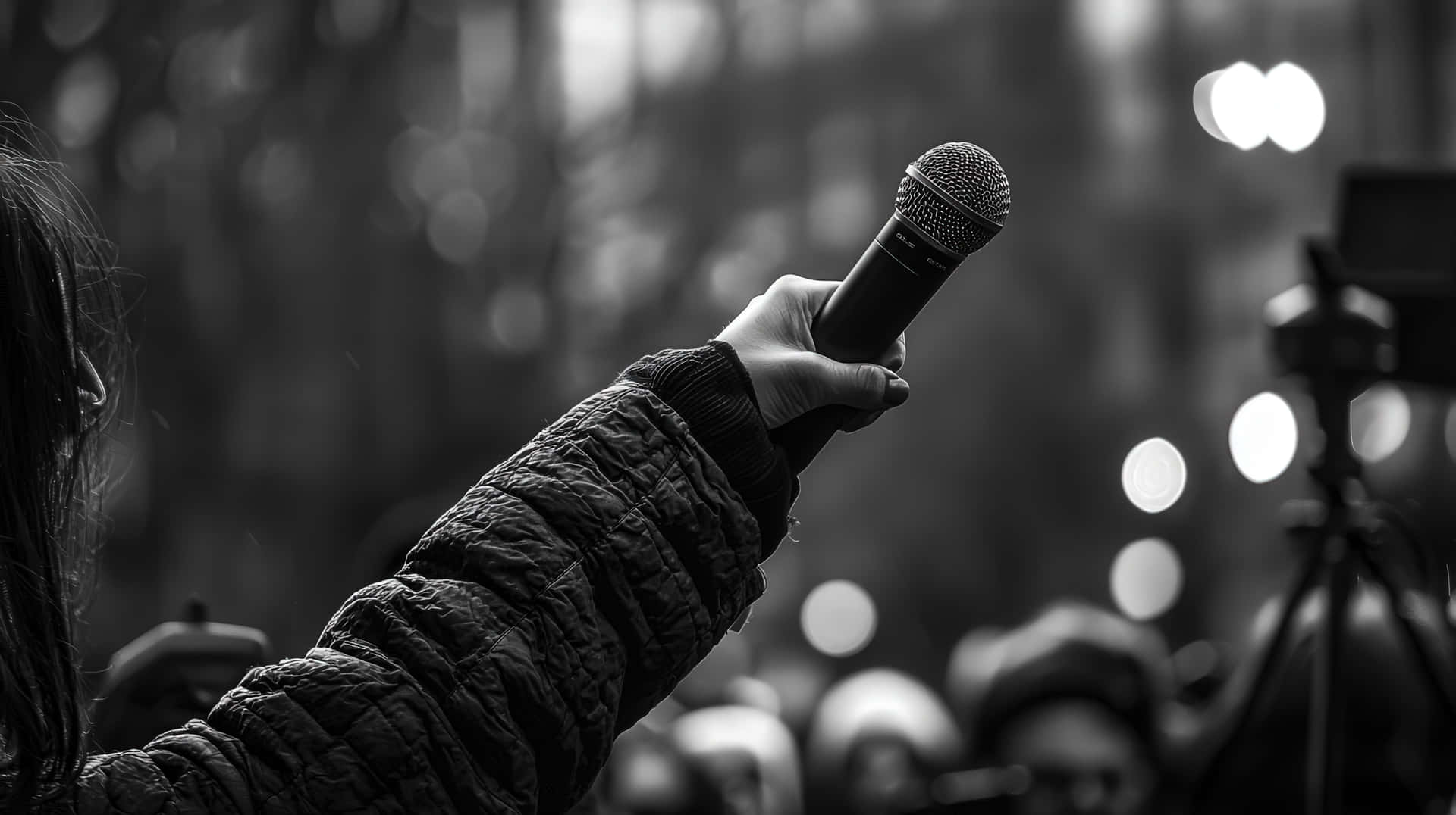 Black and White Protest Scene with a Hand Holding a Microphone for Public Speech 壁紙 — 一隻穿著絎縫袖子的手在模糊的人群和明亮的散景燈光背景下舉起麥克風，黑白影像。