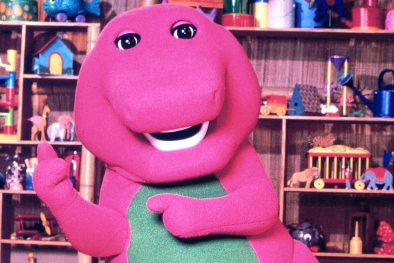 barney 桌布 壁紙 — 一隻穿著綠色背心的開心粉紅色恐龍手偶，在一個充滿色彩繽紛玩具的書架前微笑並做手勢。
