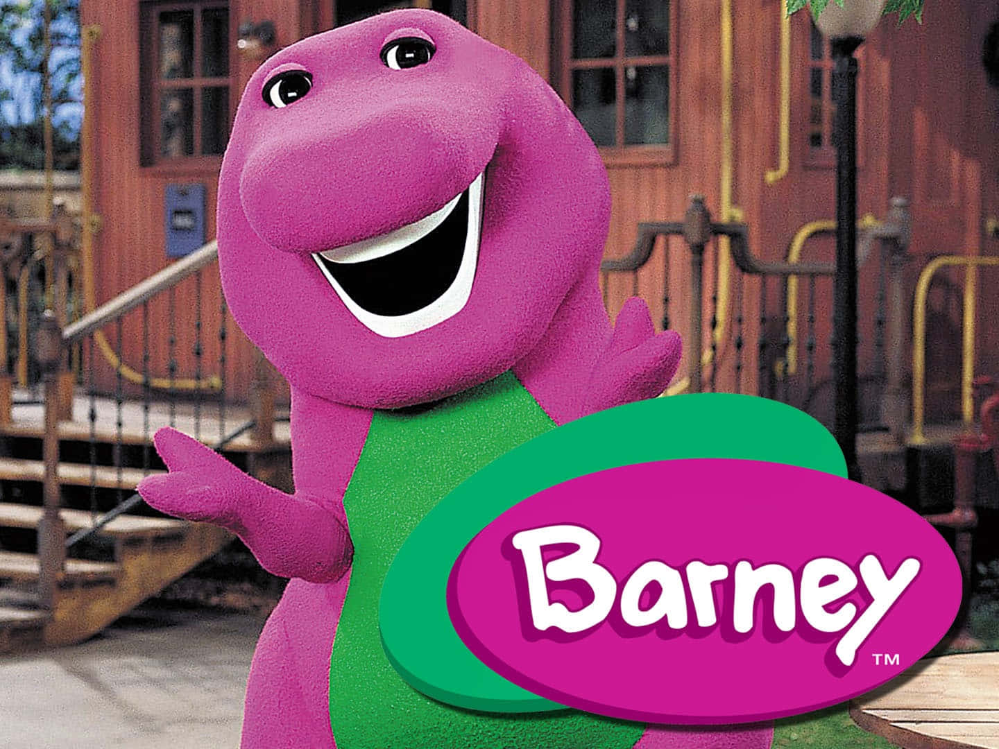 barney 桌布 壁紙 — 一隻穿著綠色襯衫的快樂紫色恐龍在木建築前開心地揮手。