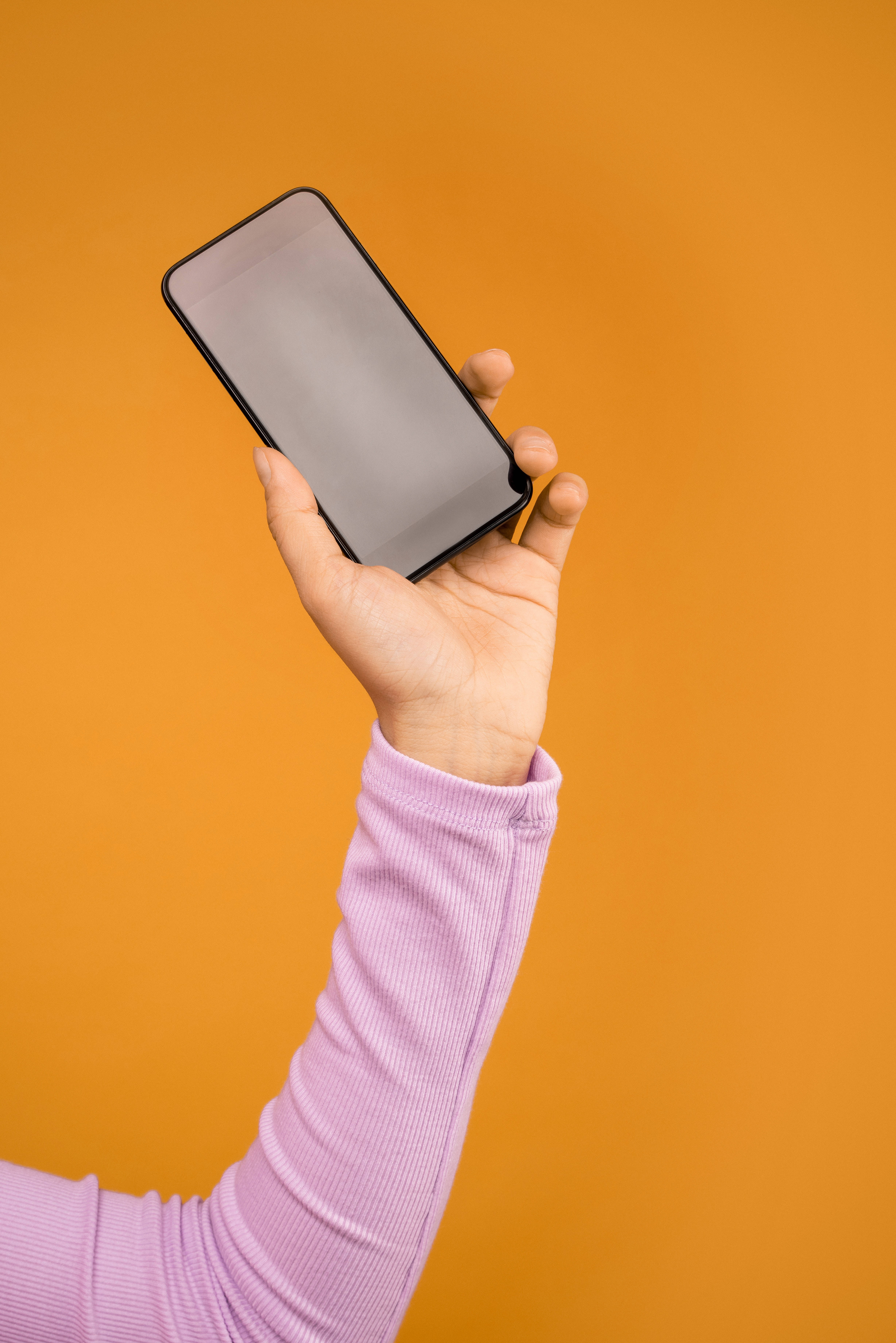 酷炫iPhone 桌布 壁紙 — 一隻穿著薰衣草色袖子的手，在純色溫暖橙色背景前拿著一支黑色智慧型手機。