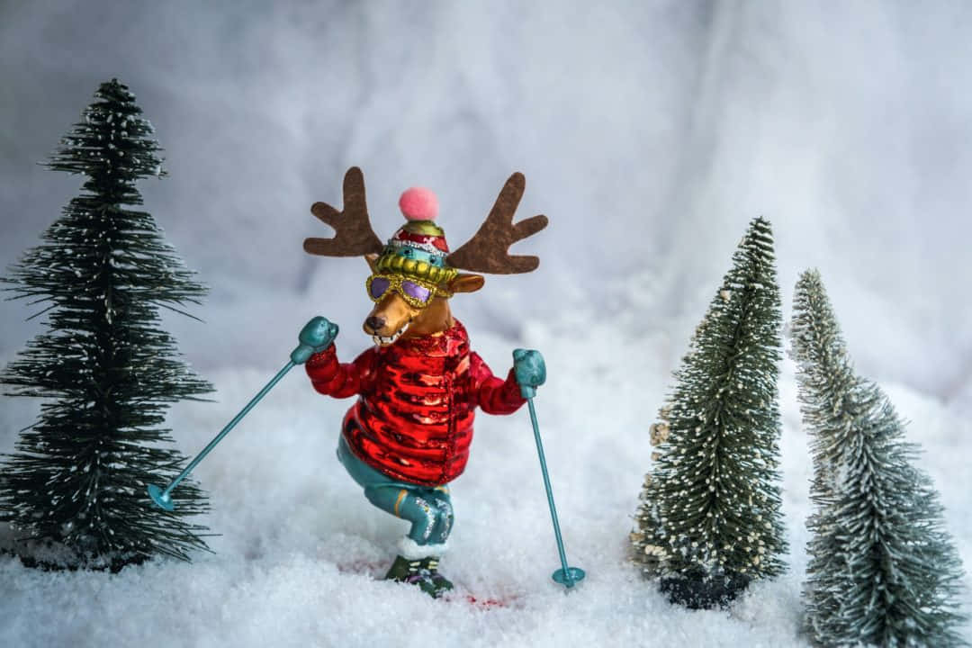 鹿 桌布 壁紙 — 一隻節日馴鹿公仔，身穿紅色亮片外套和護目鏡，在雪地中滑雪，兩旁有迷你松樹。