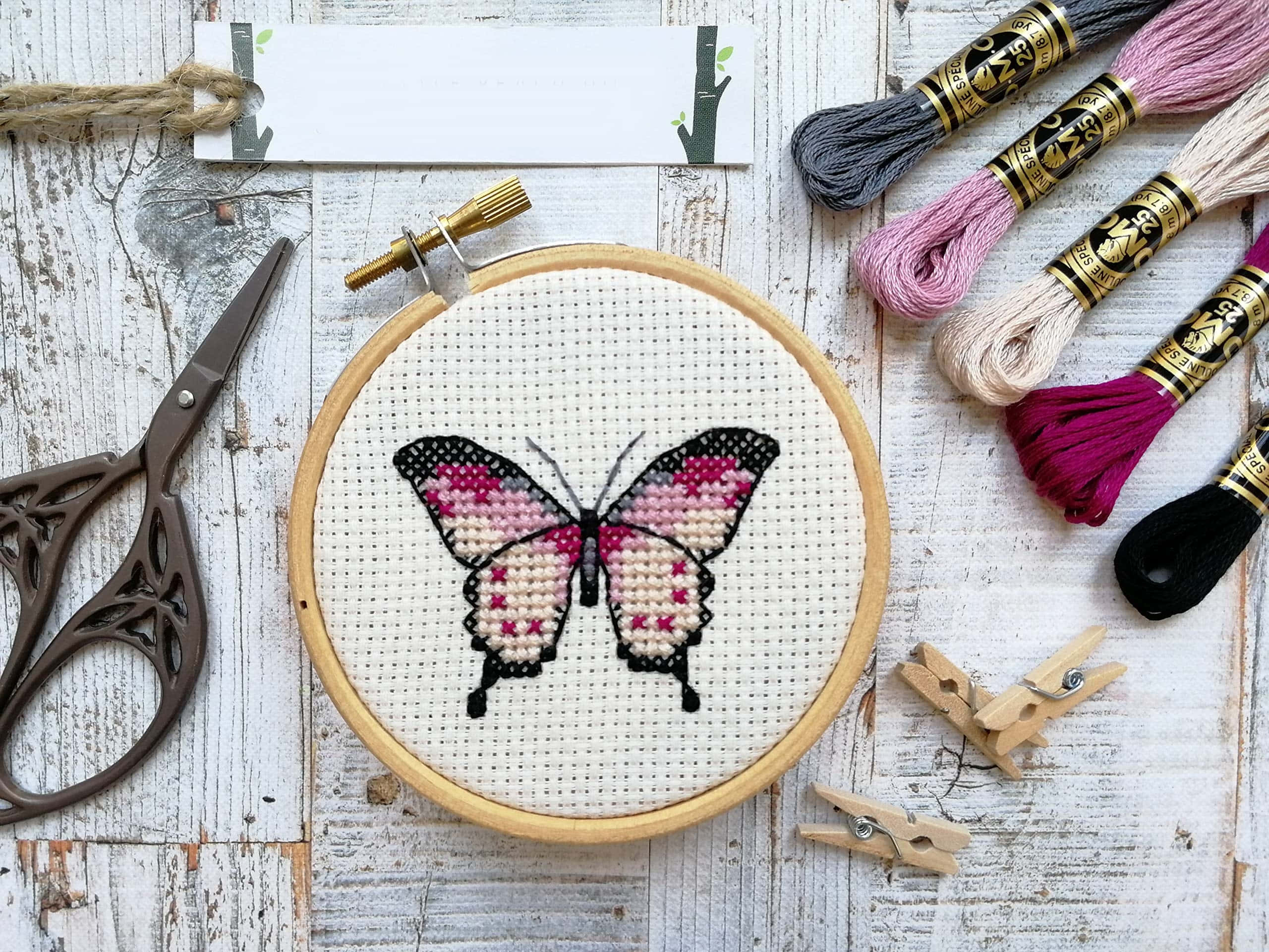 Butterfly Stitch 桌布 壁紙 — 一隻粉紅色和黑色的十字繡蝴蝶放在刺繡框內，周圍有繡花線和復古剪刀，放在風化的木質表面上。