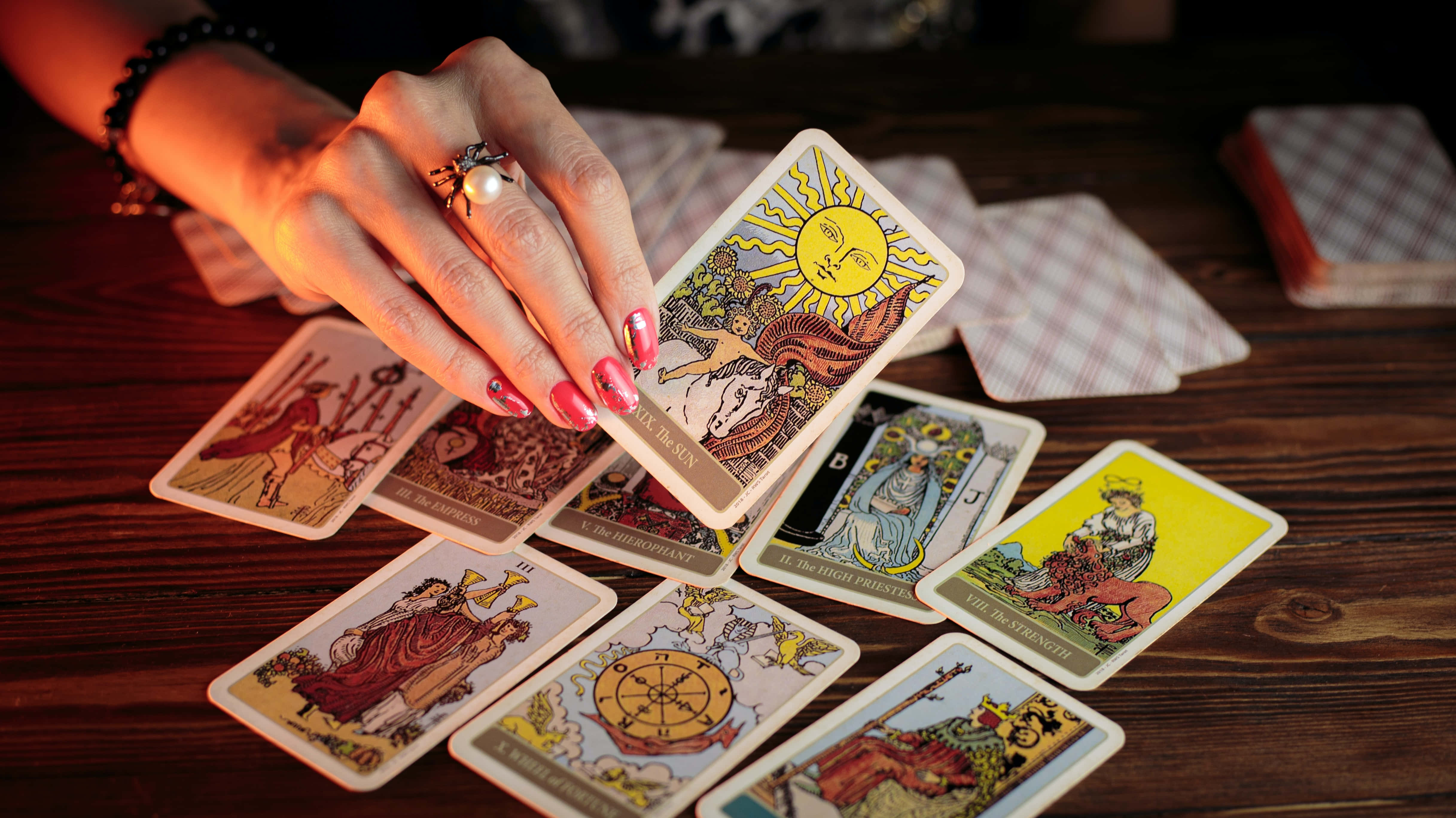 Tarot Card 桌布 壁紙 — 一隻粉紅色指甲的手在深色木桌上，於神秘卡牌攤開的上方舉起太陽塔羅牌。