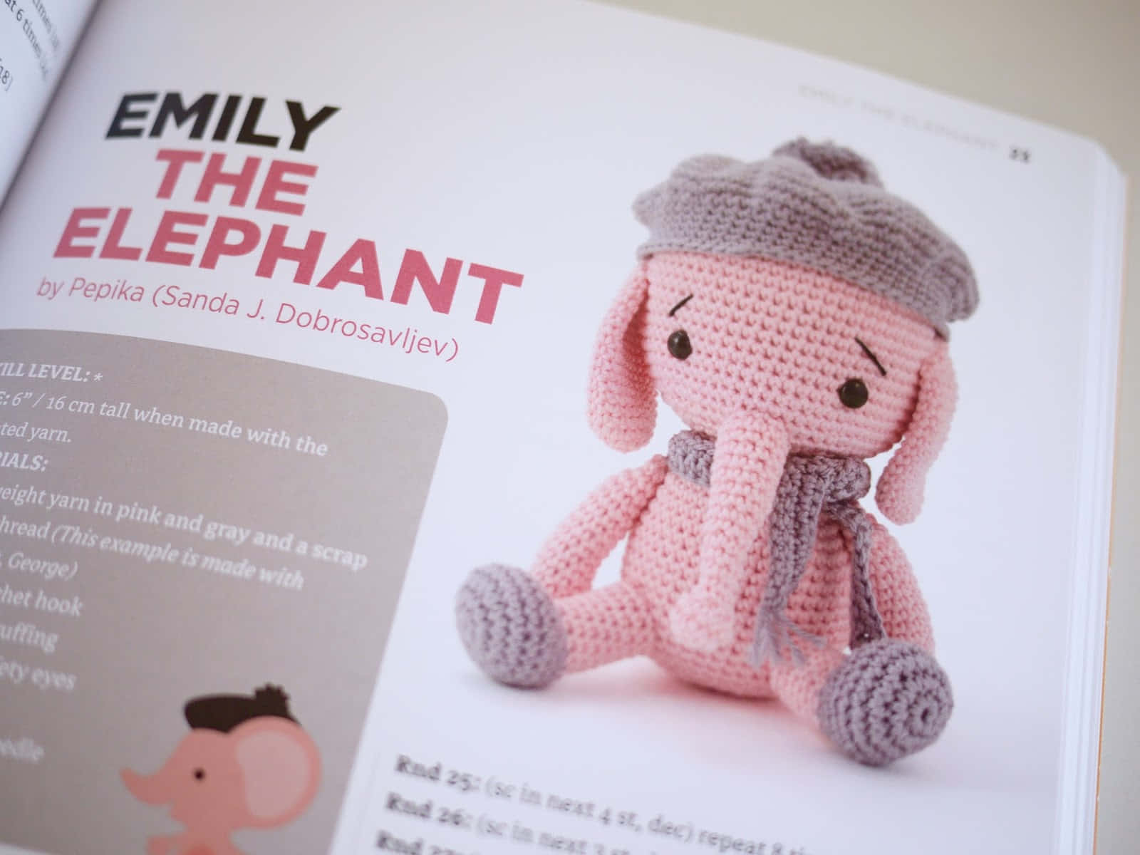 Emily Elephant 桌布 壁紙 — 一隻粉紅色鉤針大象戴著灰色帽子和圍巾，坐在一本名為《Emily the Elephant》的手工書頁旁邊。
