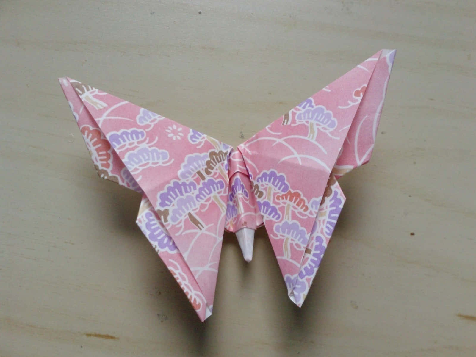 Butterfly Origami 桌布 壁紙 — 一隻精緻的粉紅色摺紙蝴蝶，帶有紫色樹木圖案，停放在淺色木質表面上。