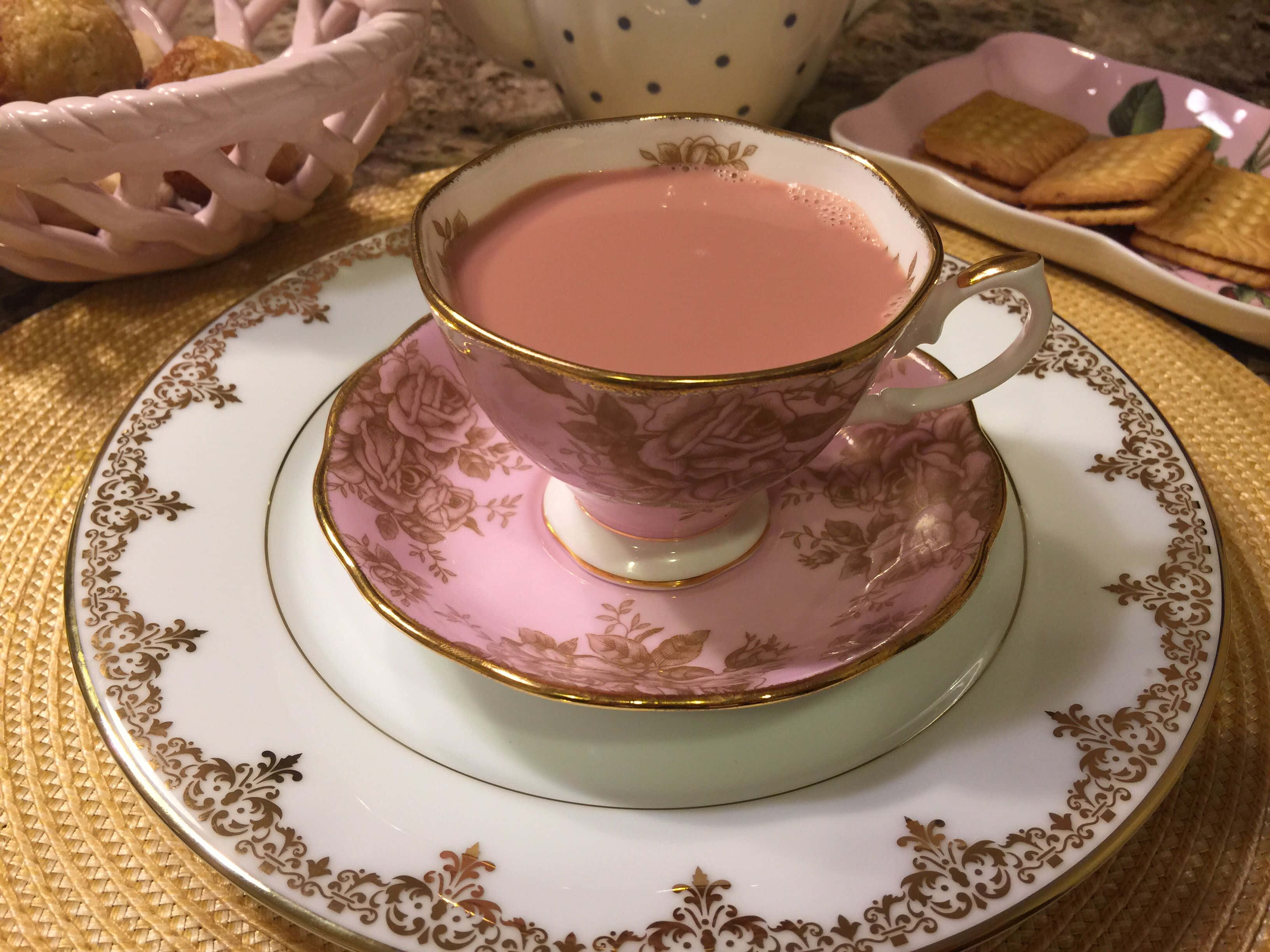 Pink Tea 桌布 壁紙 — 一隻精緻的粉紅色茶杯，帶有金色邊緣和玫瑰圖案，放在配套的茶碟上，再置於華麗的金邊盤子上，周圍有餅乾和一個編織籃。