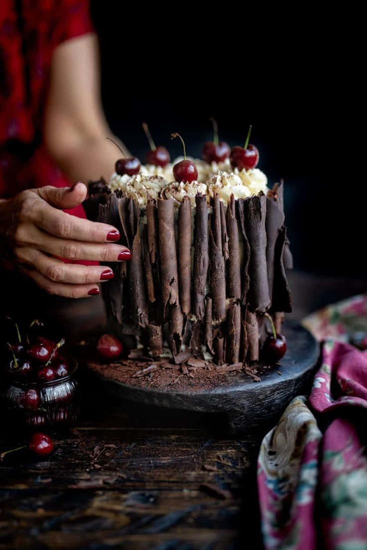 Black Forest Cake 桌布 壁紙 — 一隻紅指甲的手在深色鄉村背景下輕輕拿著一塊裝飾有櫻桃和奶油的頹廢巧克力蛋糕。