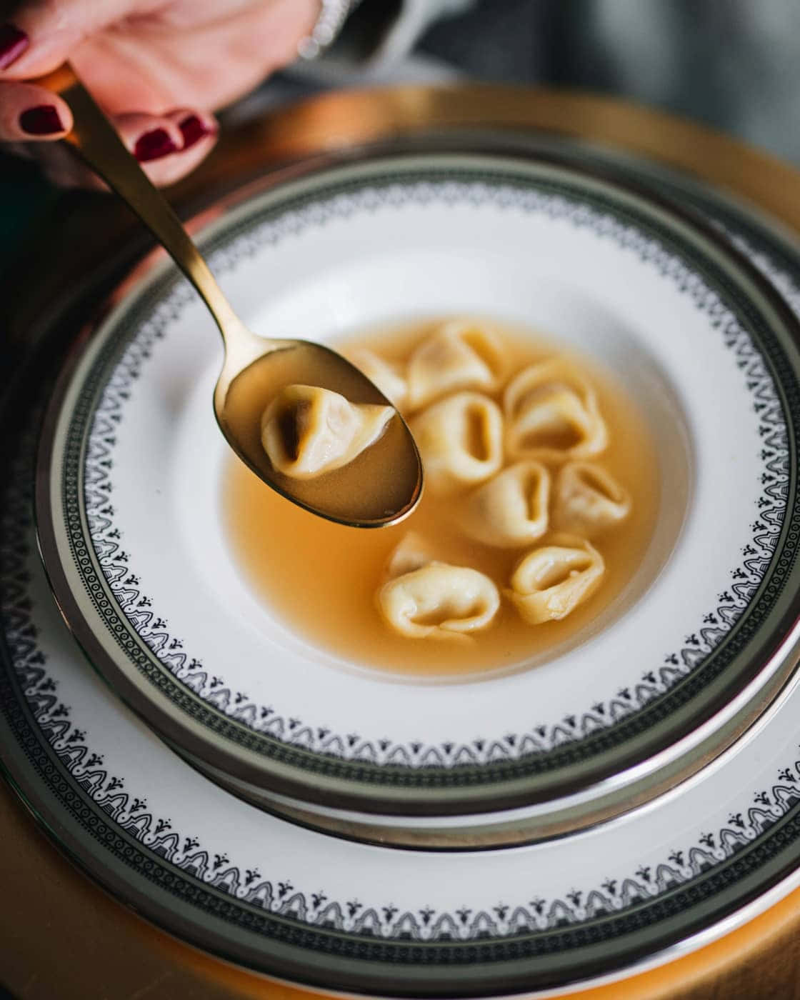 Food Photography 桌布 壁紙 — 一隻紅指甲的手從裝飾盤上的清湯碗中，用金色湯匙舀起一個義大利餃。