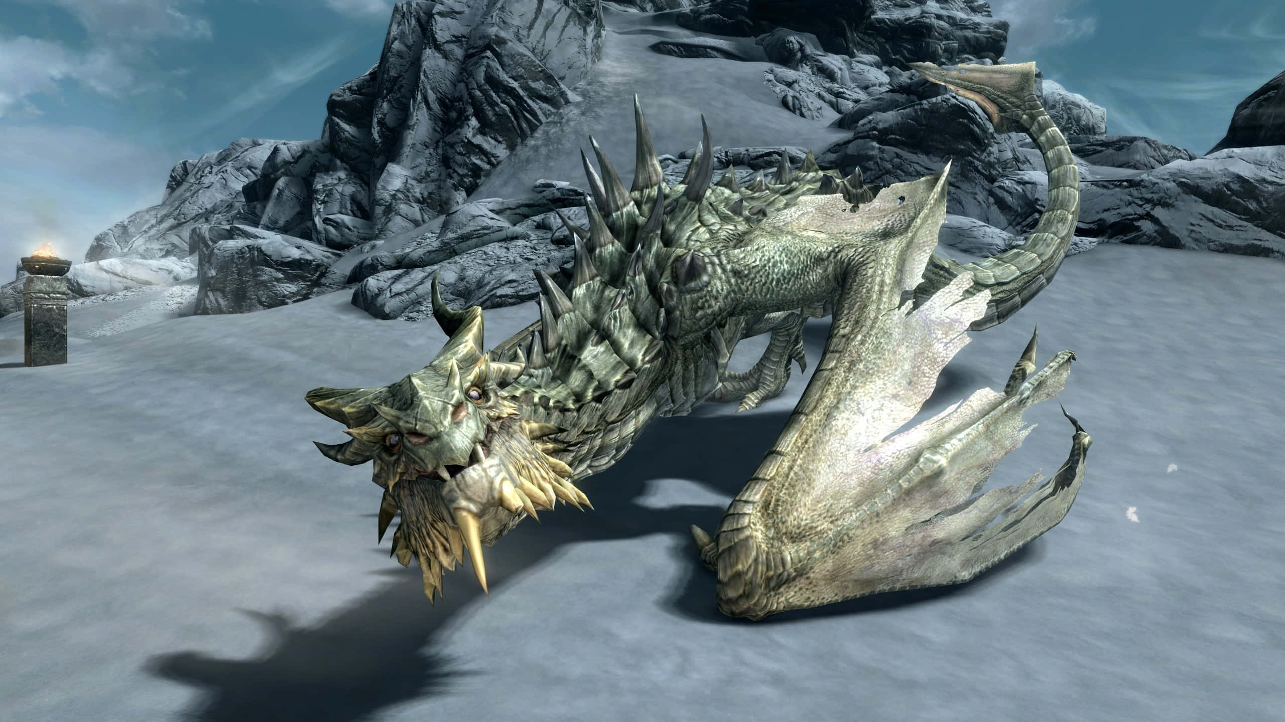 Skyrim Paarthurnax 桌布 壁紙 — 一隻細節豐富的3D渲染龍，帶有尖刺鱗片和金色鬃毛，在雪地上休息，旁邊是崎嶇的岩石，天空多雲。