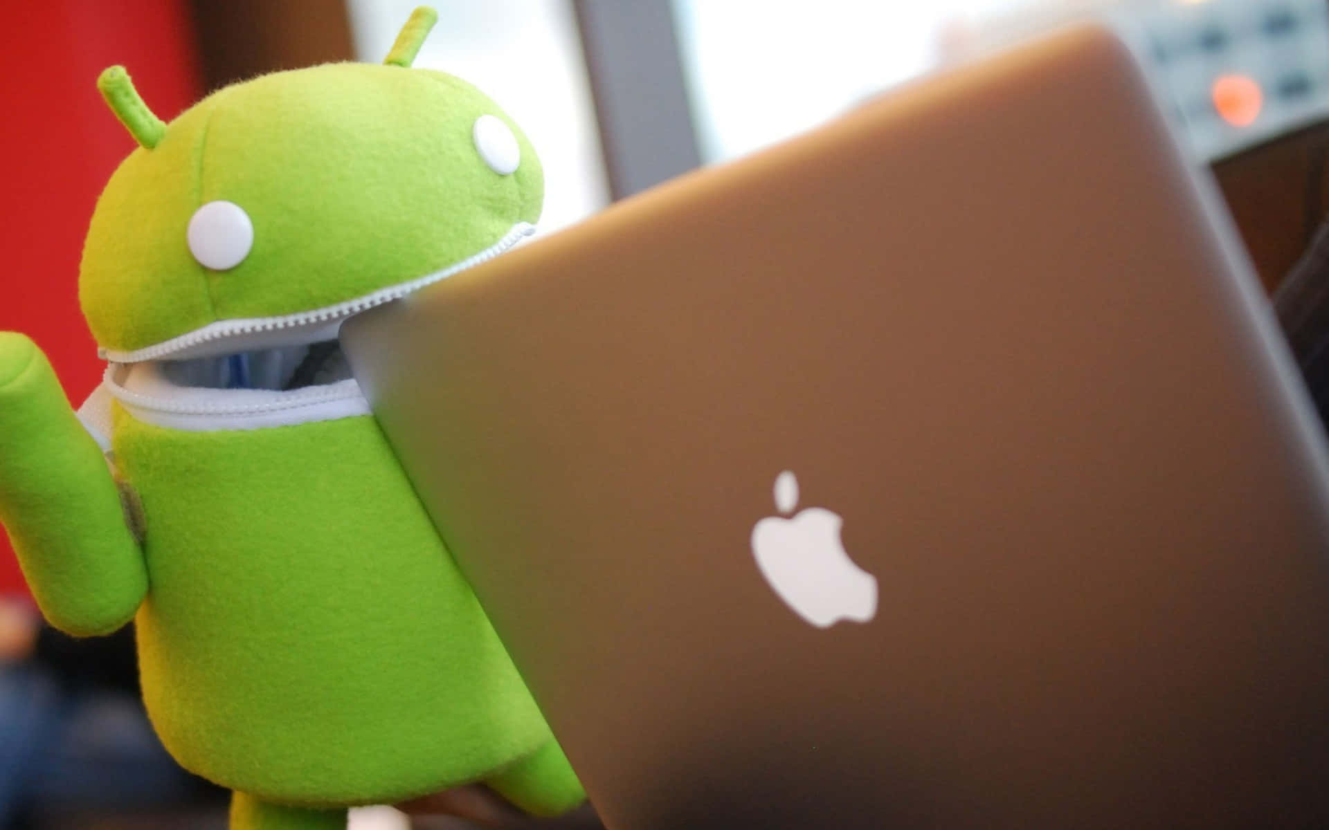 蘋果標誌 桌布 壁紙 — 一隻綠色Android絨毛娃娃從帶有Apple標誌的銀色筆記型電腦後面探出頭來。