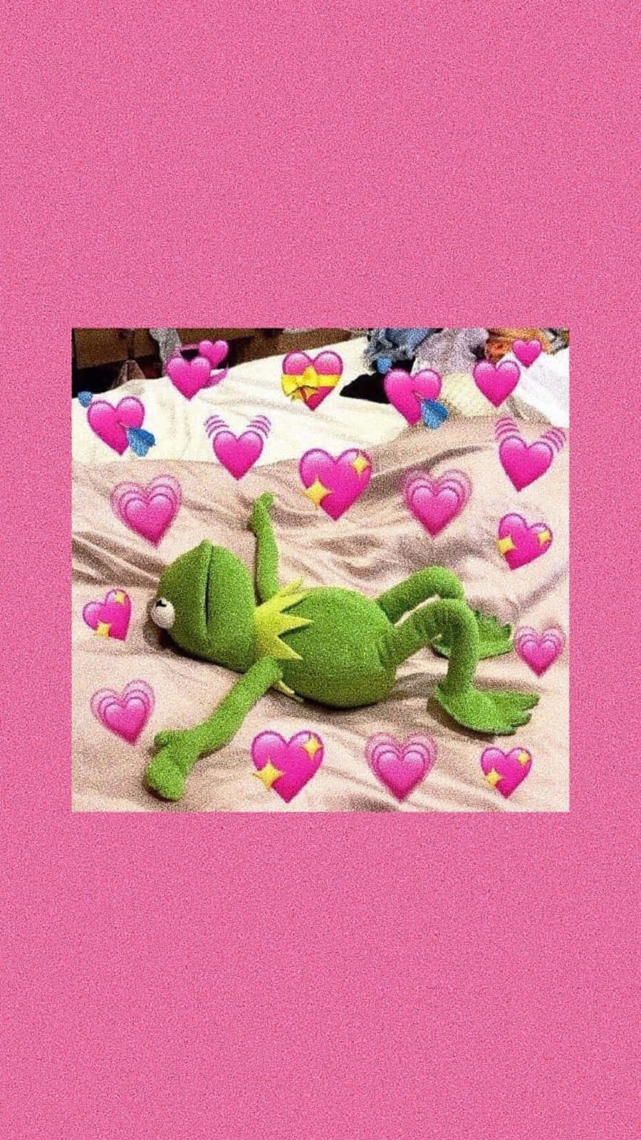 可愛的青蛙 桌布 壁紙 — 一隻綠色Kermit the Frog絨毛玩具放鬆地躺在床上，周圍漂浮著粉紅色愛心表情符號，背景為純粉紅色。