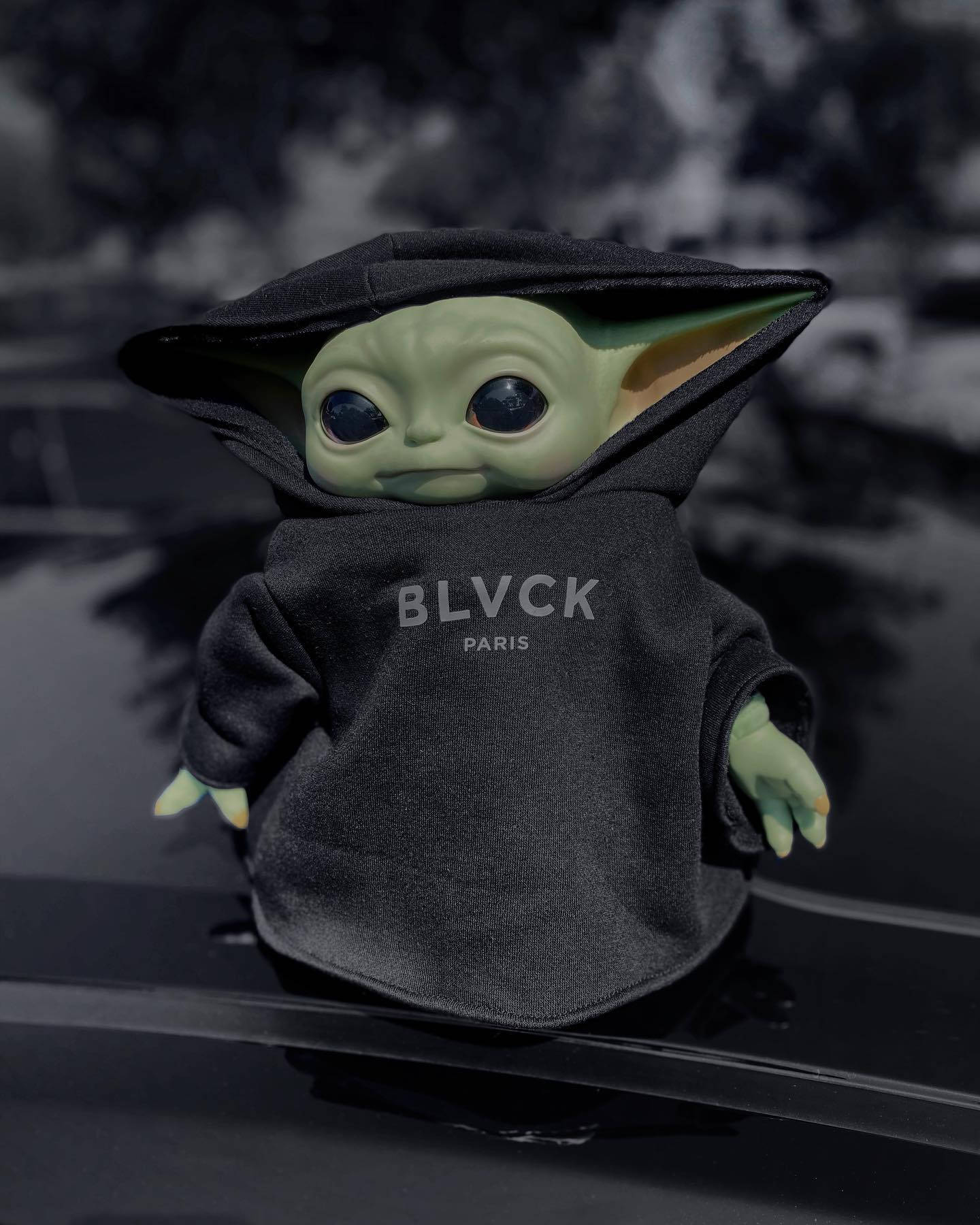 Baby Yoda絨毛玩偶穿著黑色連帽上衣，印有BLVCK PARIS字樣，坐在汽車引擎蓋上。