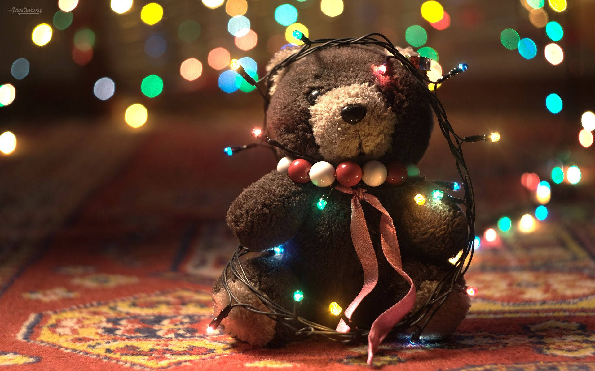 Cute Bear 桌布 壁紙 — 一隻舒適的棕色泰迪熊，點綴著閃爍的仙女燈和粉紅色絲帶，坐在圖案地毯上，背景是色彩繽紛的燈光散景。
