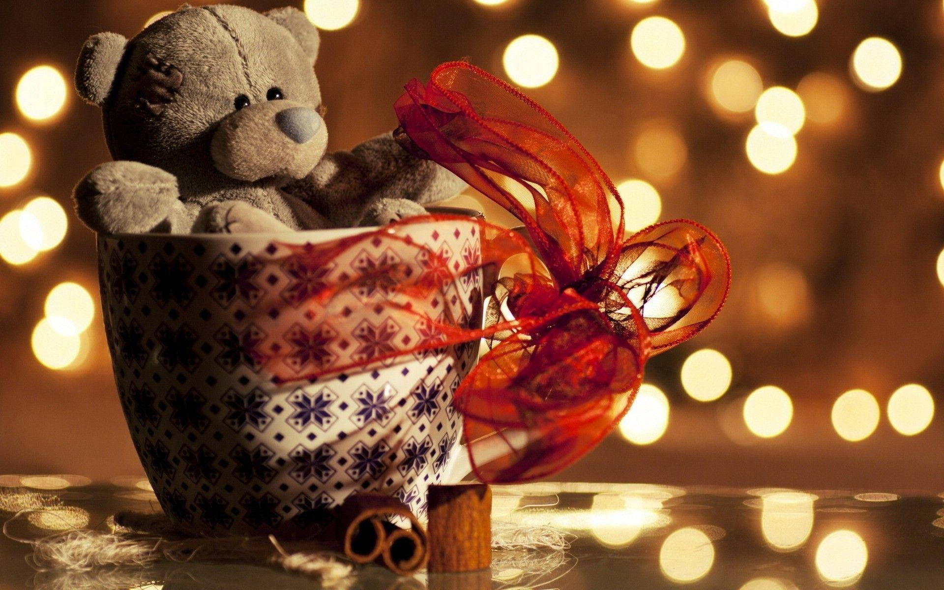 Teddy Bear 桌布 壁紙 — 一隻舒適的灰色泰迪熊坐在有圖案的杯子裡，旁邊有肉桂棒和紅色絲帶，背景是溫暖的散景燈光。