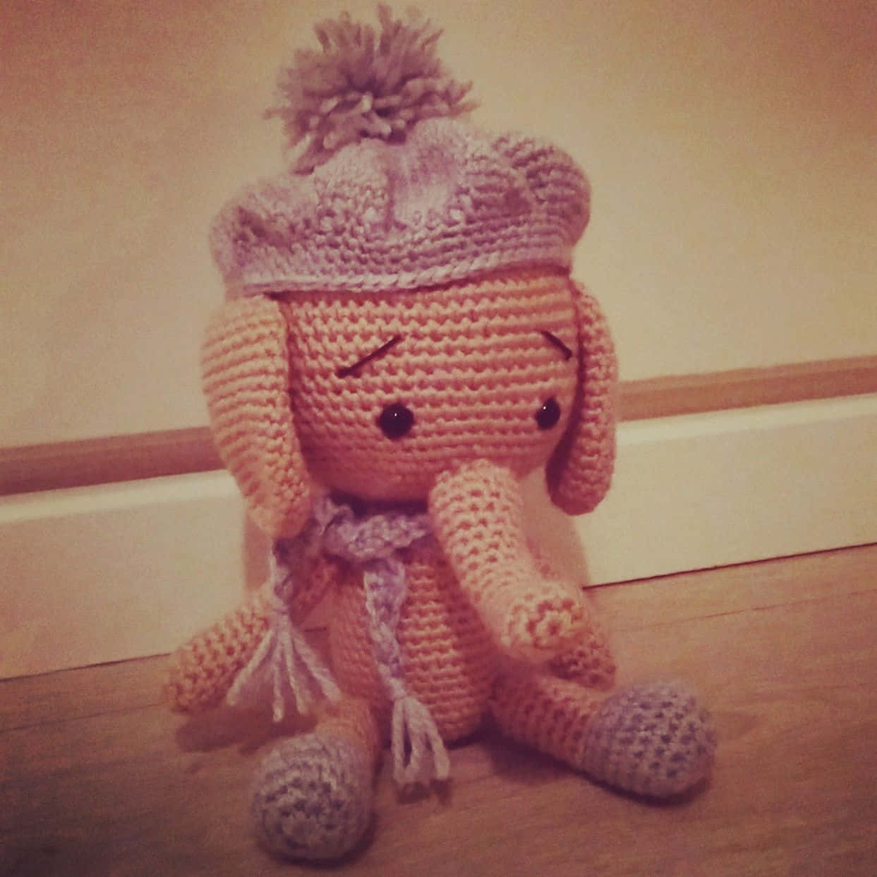 Emily Elephant 桌布 壁紙 — 一隻舒適的鉤針編織大象絨毛玩具，戴著有絨球的針織帽，坐在踢腳板附近的木地板上。