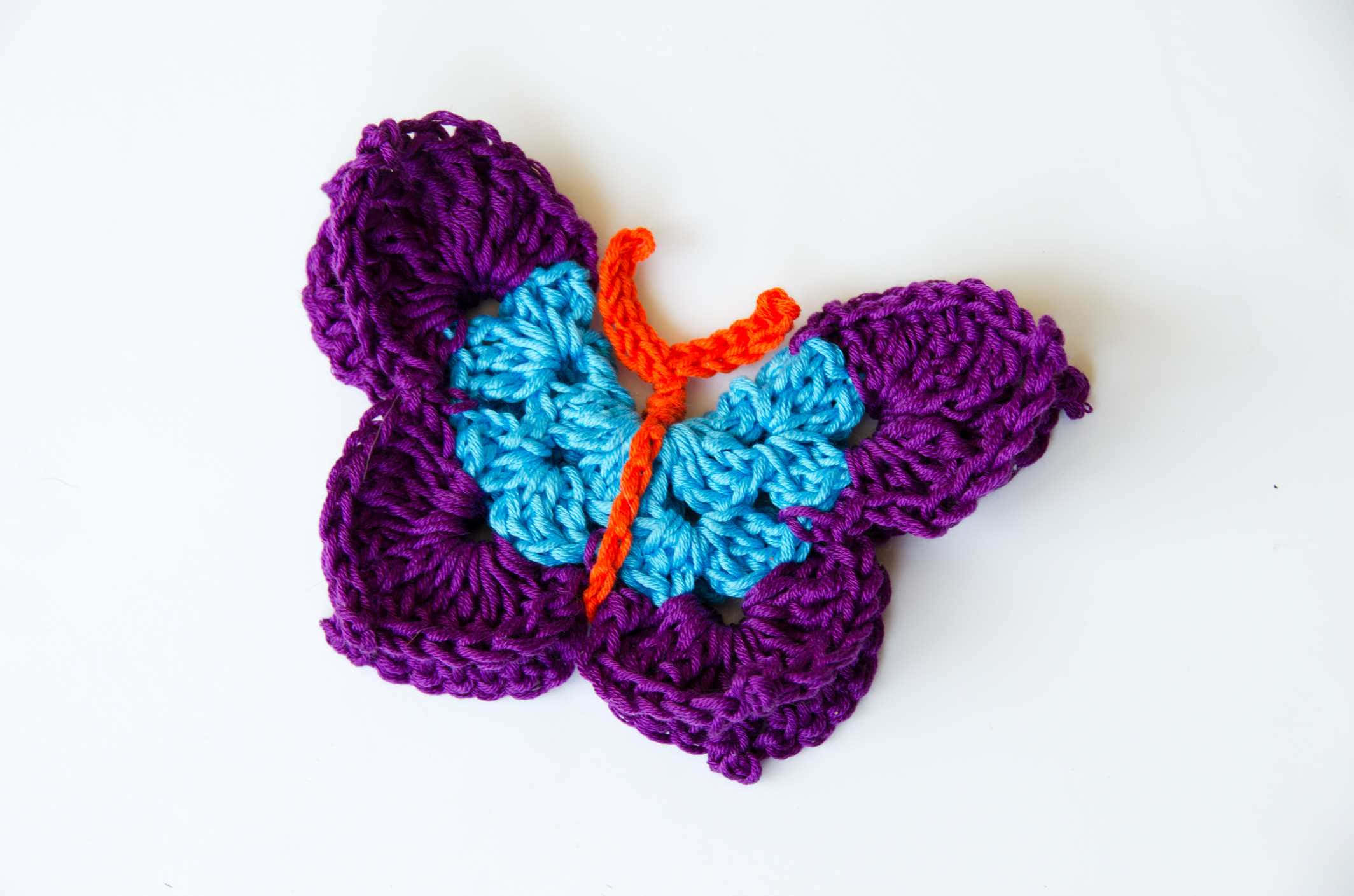Butterfly Stitch 桌布 壁紙 — 一隻色彩繽紛的鉤針編織蝴蝶，帶有紫色翅膀和藍色中心，放在純白色表面上。